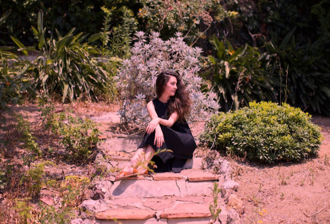Woman with long curly brown hair sitting on stone garden steps surrounded by foliage