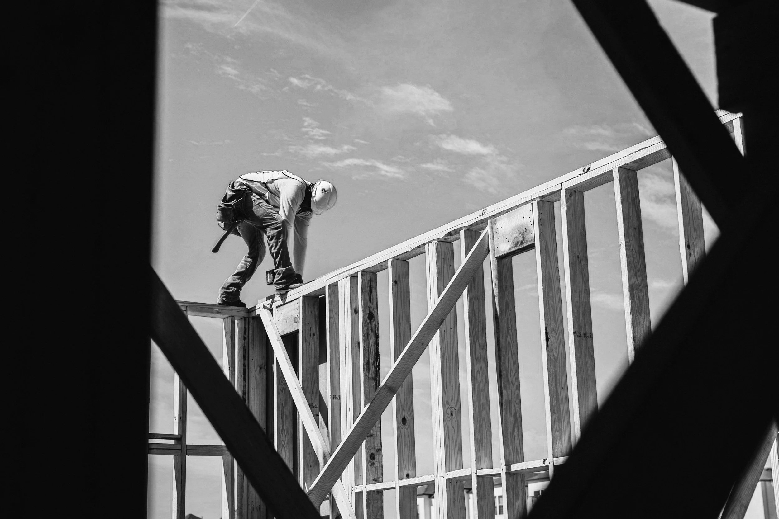A construction worker wearing a helmet and work clothes is working on the wooden frame of a building under construction, seen through a framing opening.