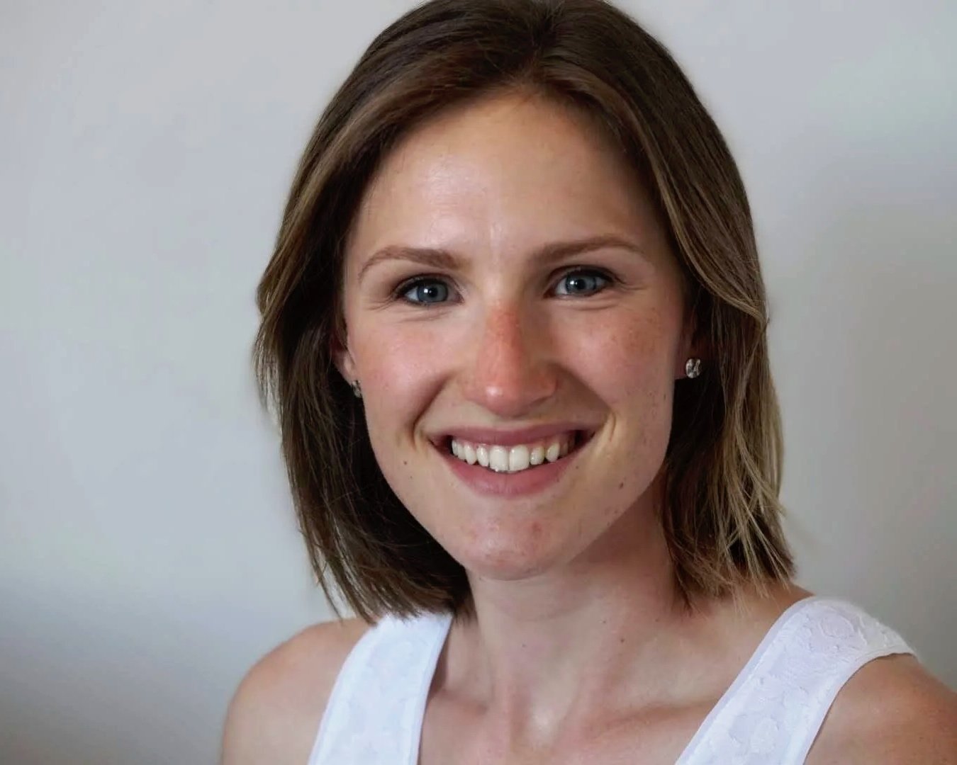 Close-up of a smiling woman with short brown hair, blue eyes, wearing diamond stud earrings and a white sleeveless top, against a plain light background.