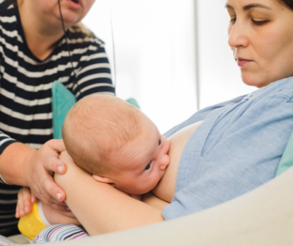 A woman breastfeeding a baby while a person in striped shirt holds the baby's head.