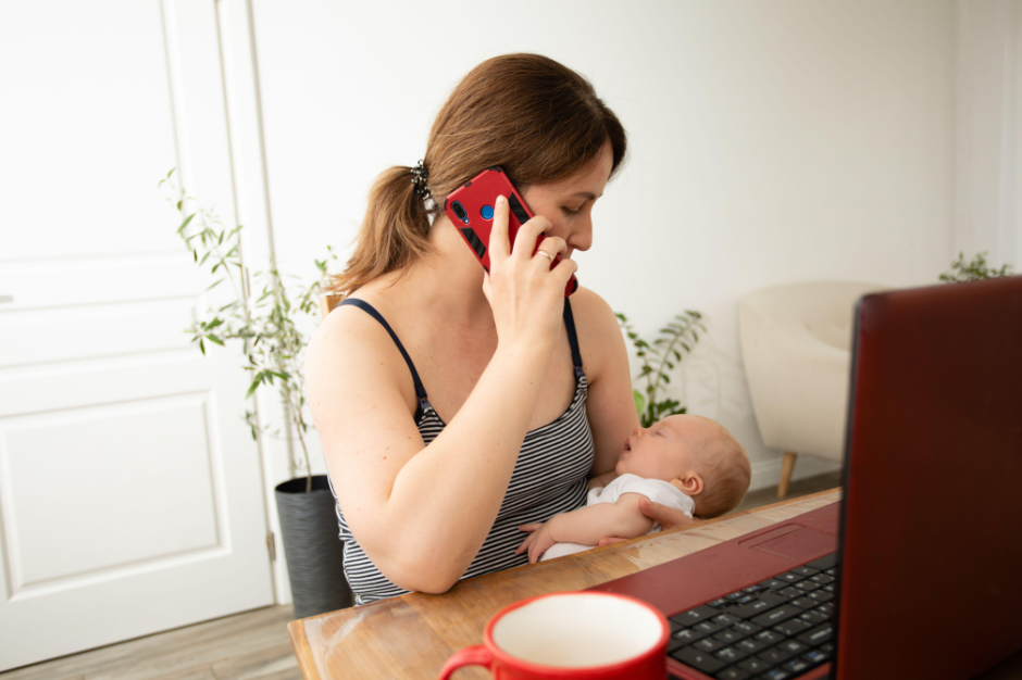 A woman holding a baby at a table while talking on a phone, with a laptop and a mug in front of her, in a bright room with plants and a white door in the background.