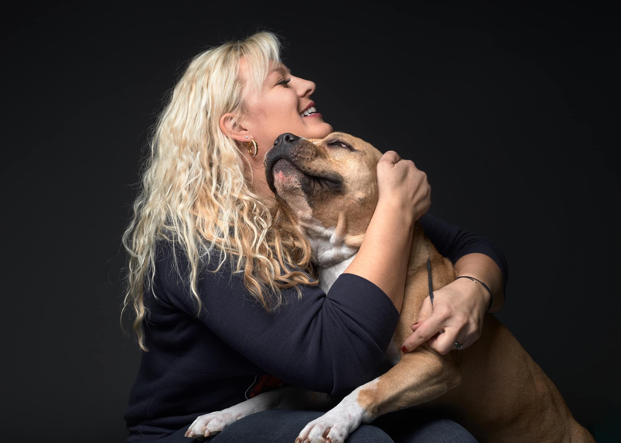 A woman with blond, curly hair hugging a large brown and white dog, both appearing happy and affectionate against a dark background.