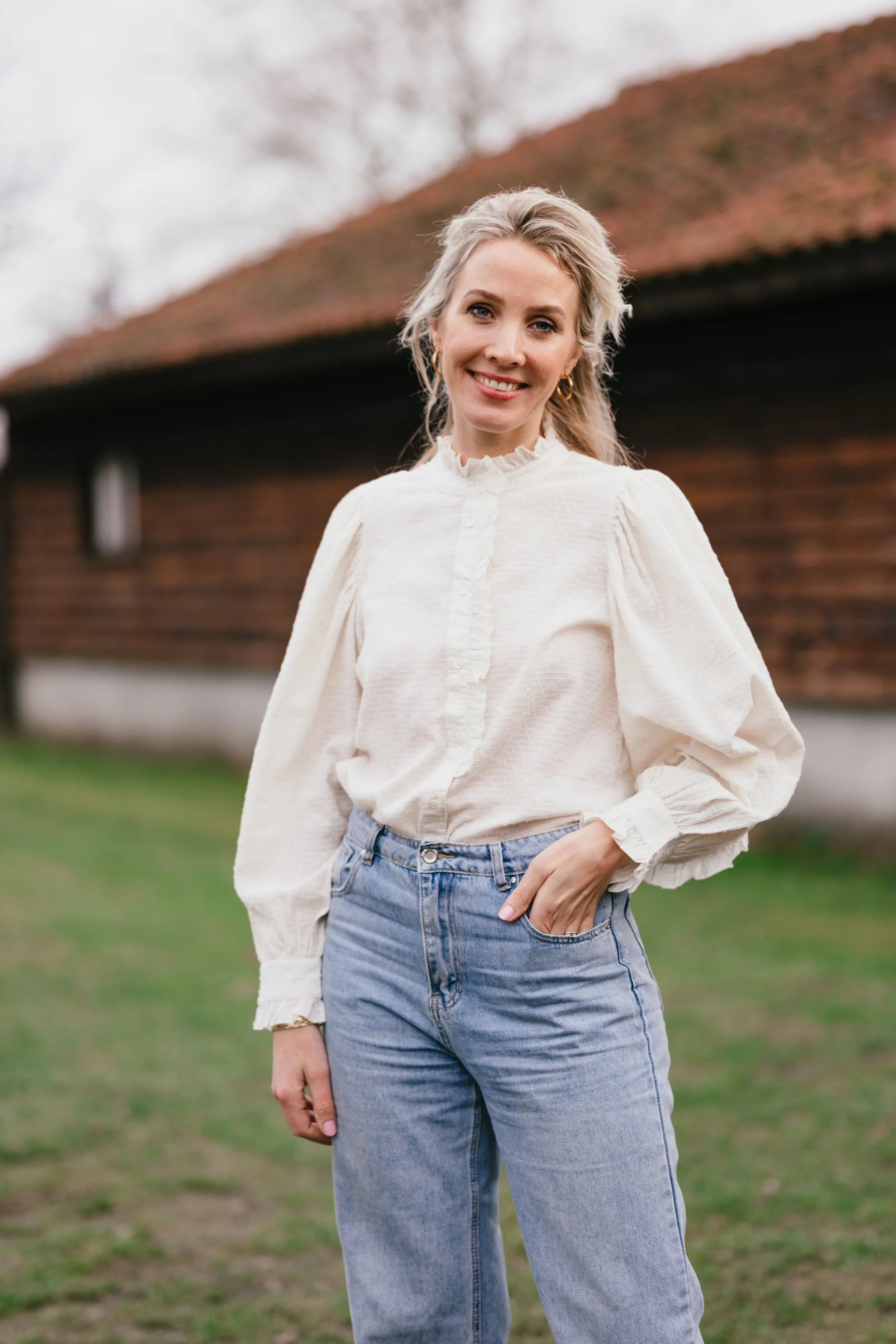 Vrouw glimlachend buiten met witte blouse en blauwe spijkerbroek, achtergrond met gras en houten gebouw.