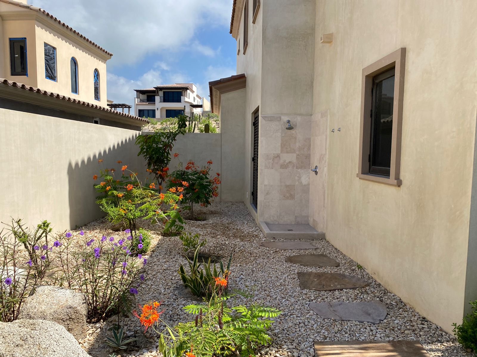 Side yard of a house with desert-style landscaping, including flowering plants, gravel, and stepping stones, with neighboring houses visible in the background under a partly cloudy sky.