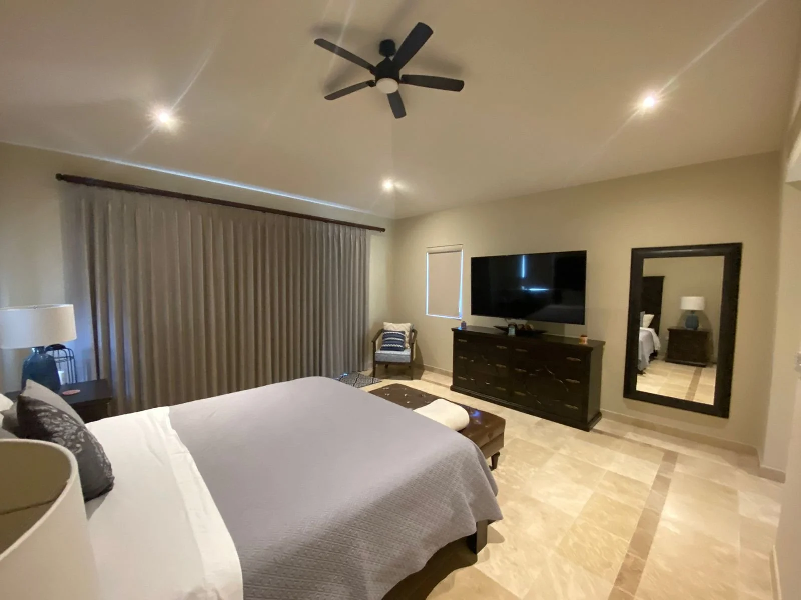 A bedroom with a large bed, a flat-screen TV on a dresser, a mirror on the wall, a ceiling fan, beige curtains, and side table with lamps.