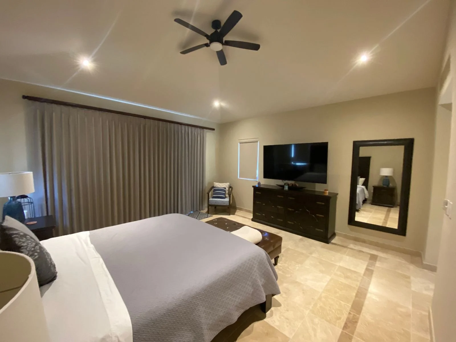 A bedroom with a large bed, nightstand, lamp, armchair, dresser with a television, a large mirror, window with closed blinds, ceiling fan, and two ceiling lights.