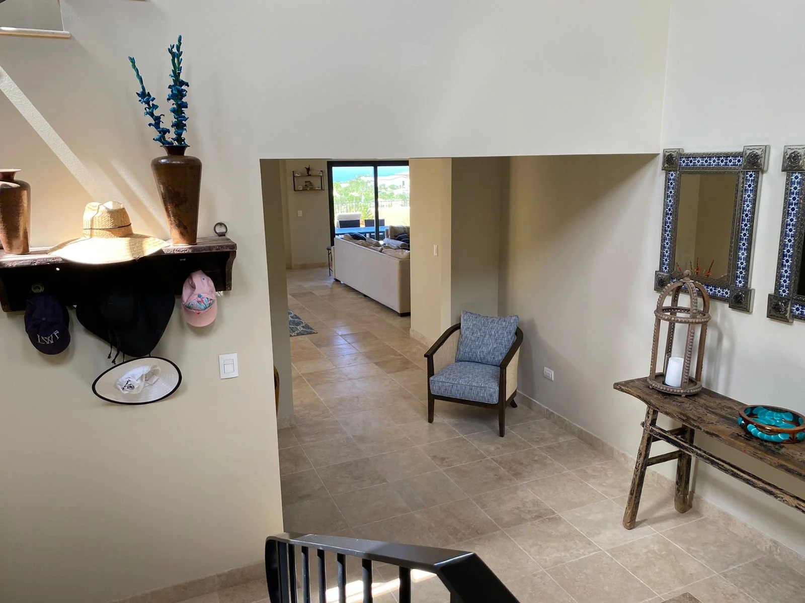 Interior view of a living room with beige tiled flooring, a wall with a rustic wooden shelf holding decorative vases and hats, a small armchair with gray upholstery, a wooden console table with a candle, and a wall mirror framed with decorative tiles