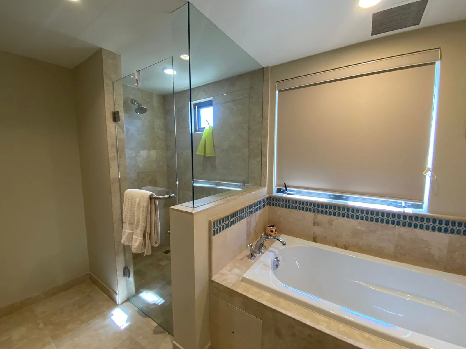 A modern bathroom with a glass-enclosed shower, a bathtub with a decorative tile border, and a window with a closed beige roller shade.