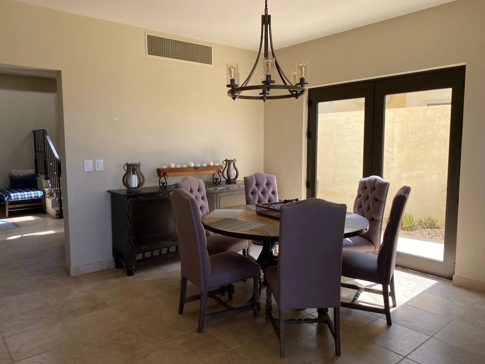 Dining room with a round wooden table, six upholstered chairs, a black chandelier, a sideboard with decorative lanterns and candles, sliding glass doors leading outside, and beige tiled flooring.