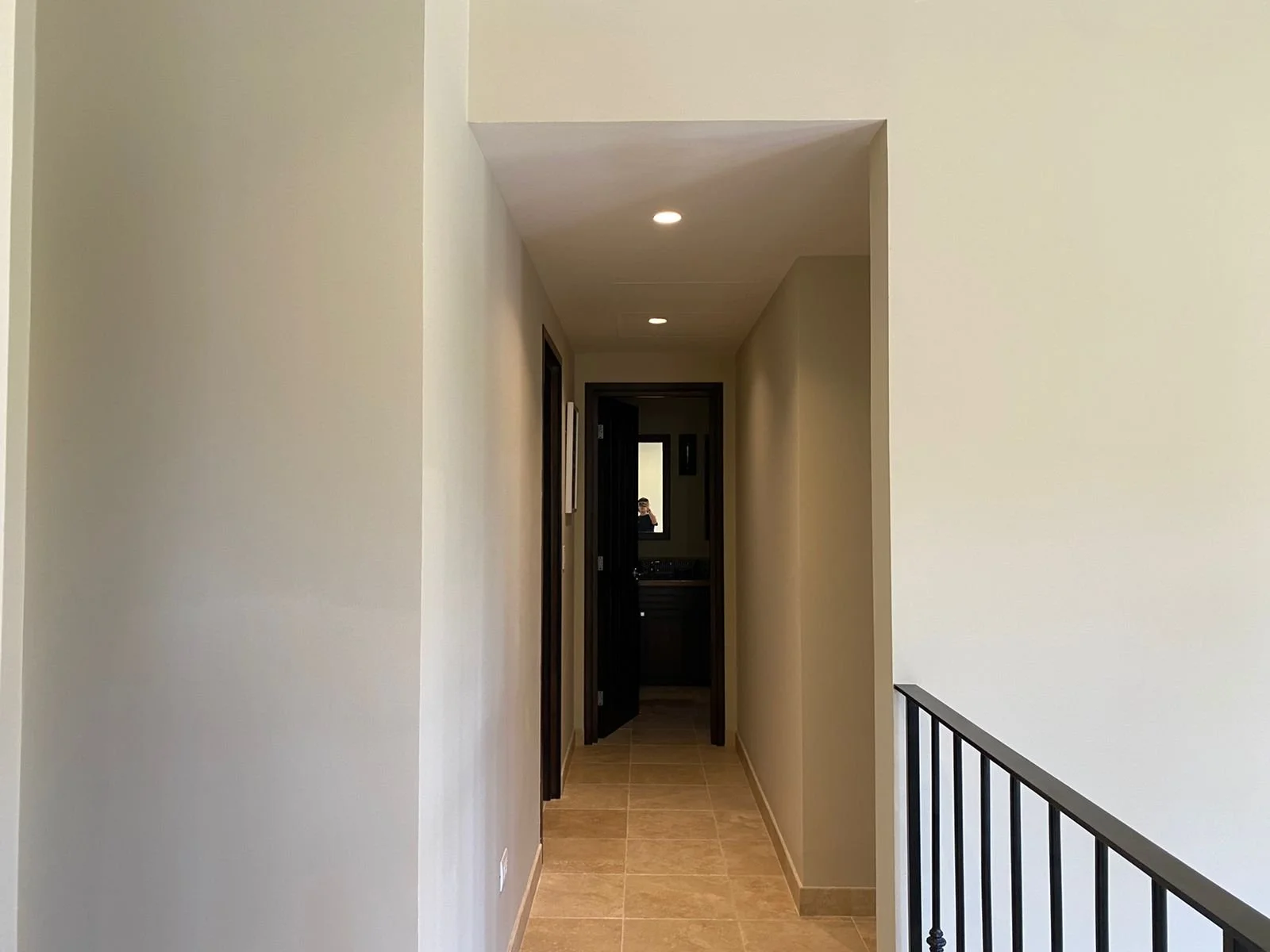 A hallway with beige tiled flooring, light-colored walls, a black door at the end, and a mirror visible through the doorway. There are recessed ceiling lights and a black railing on the right side.