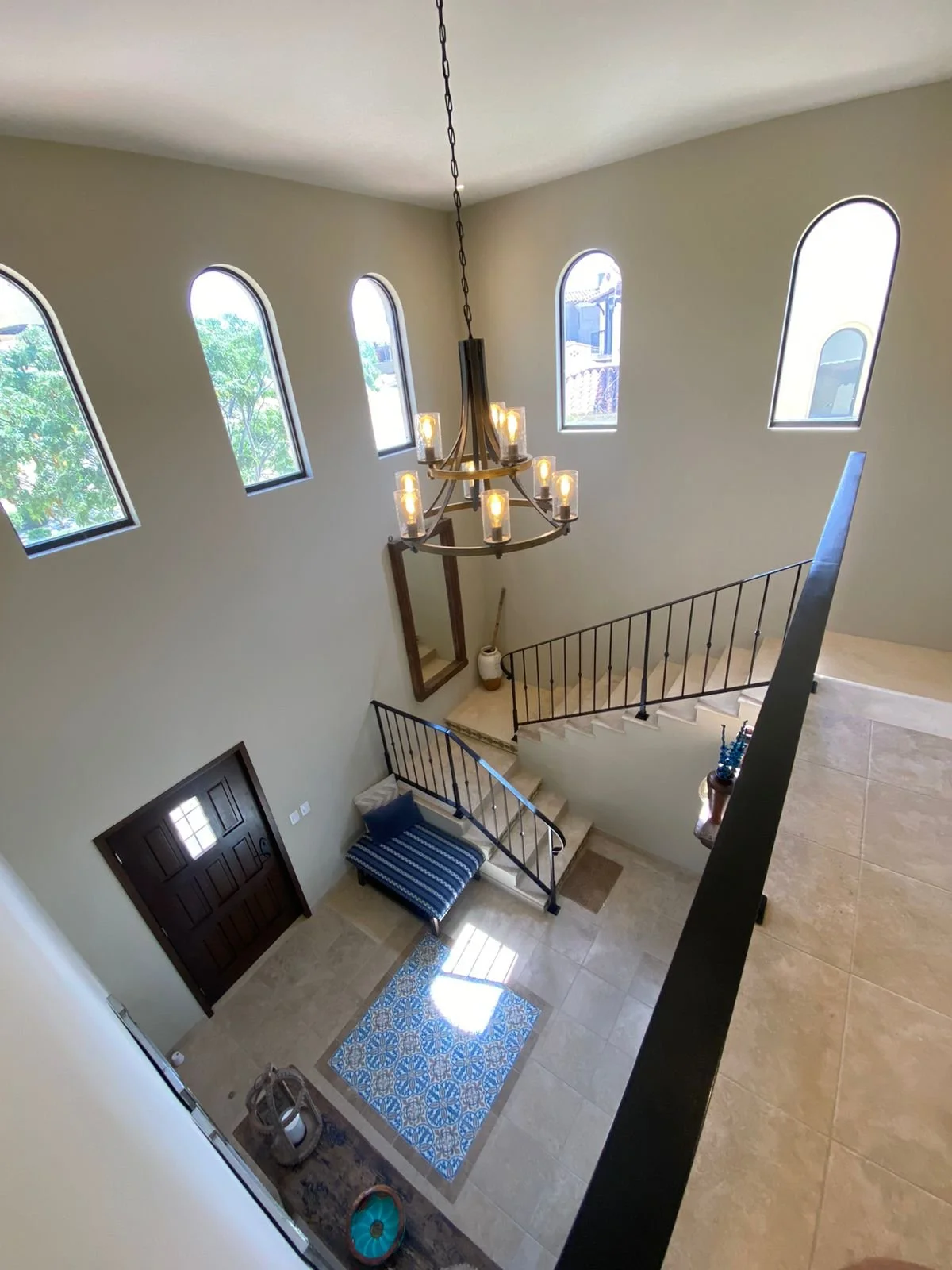 Interior view of a two-story foyer with a high ceiling, three tall arched windows, a hanging chandelier, a dark wooden front door, a striped bench with pillows, tiled flooring, and decorative items.