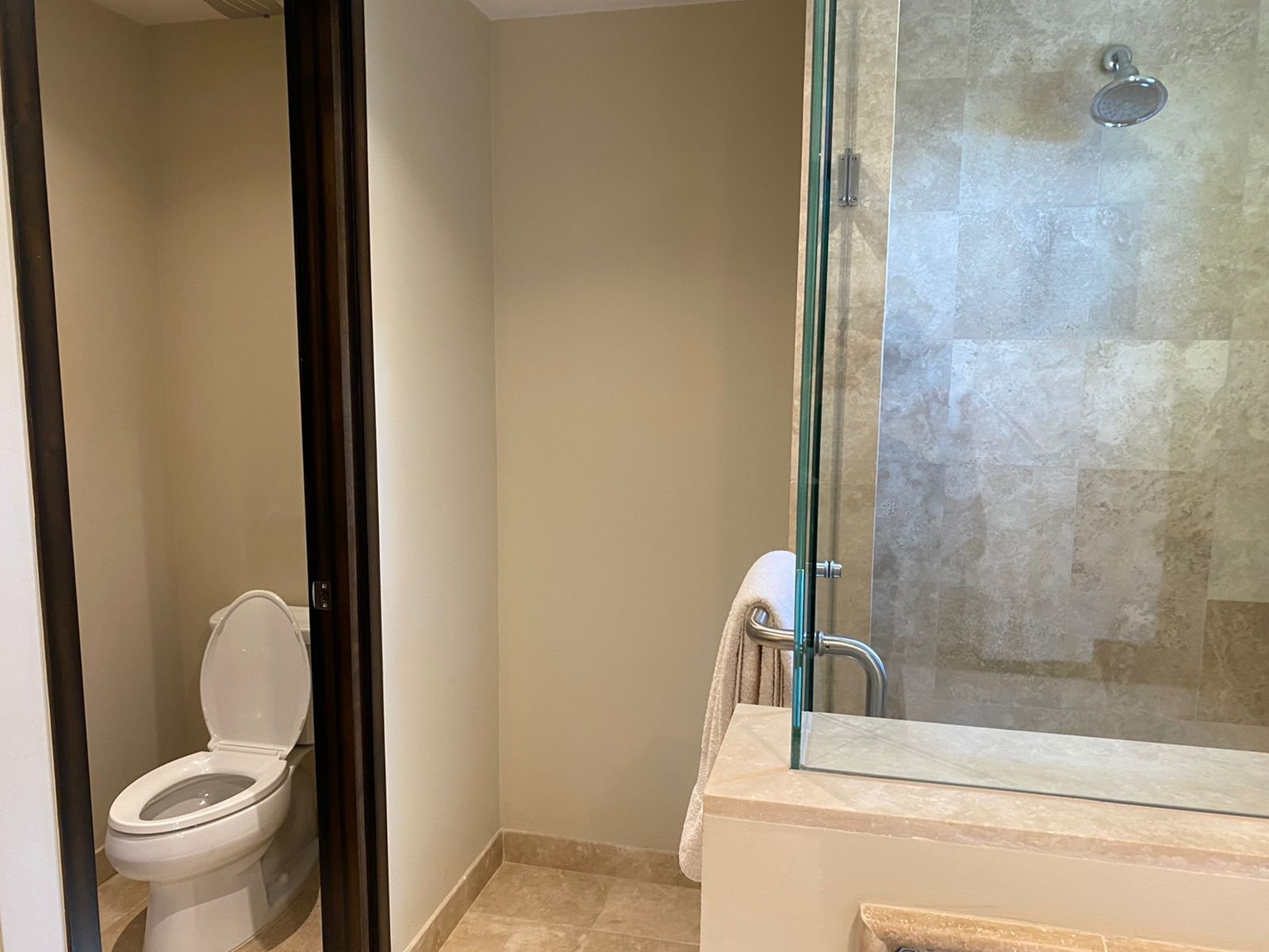 A bathroom with a toilet in a separate enclosed space and a walk-in shower with beige tiles, a glass door, and a silver shower head.
