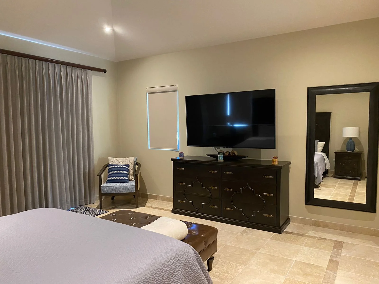 A hotel room with a bed, a chair with pillows, a large flat-screen TV on a dark wooden dresser, a large mirror on the wall, a window with closed blinds, and beige tiled flooring.