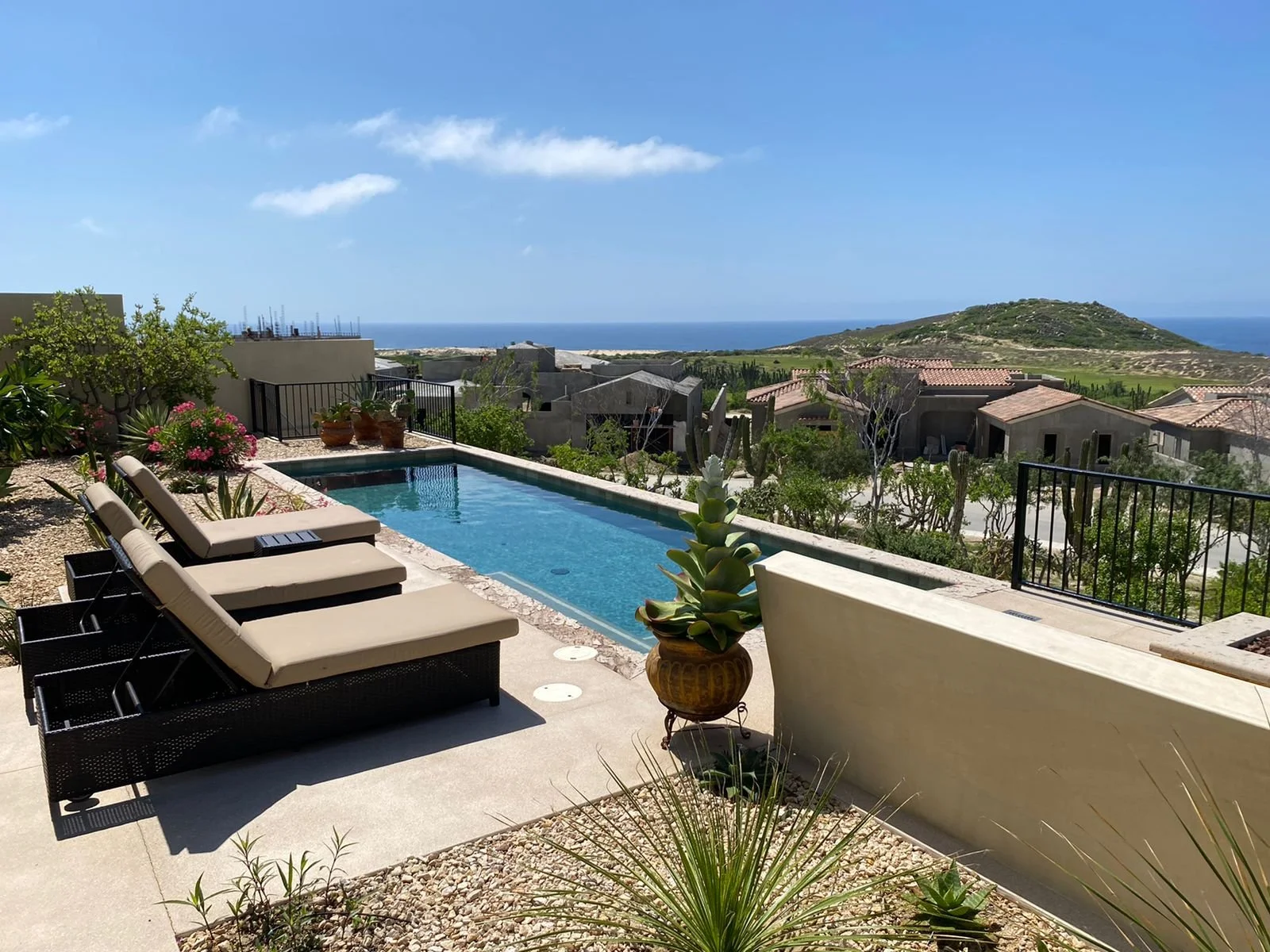 A backyard with a swimming pool, two lounge chairs, potted plants, and a view of rooftops, hills, and the ocean in the distance under a blue sky.