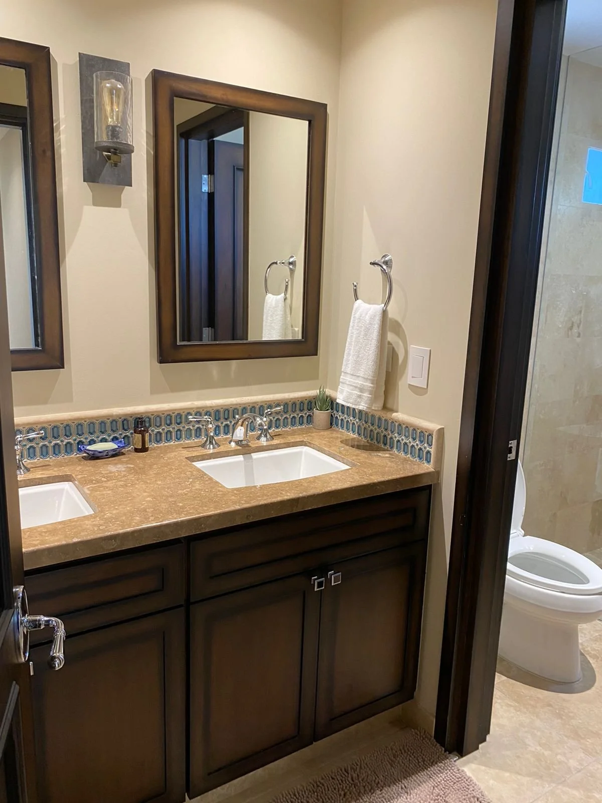 A bathroom with a double vanity featuring a beige granite countertop, two undermount sinks, dark wood cabinets, and decorative tile border behind the sinks. A mirror hangs above the vanity, with a wall-mounted light fixture beside it. There are towel