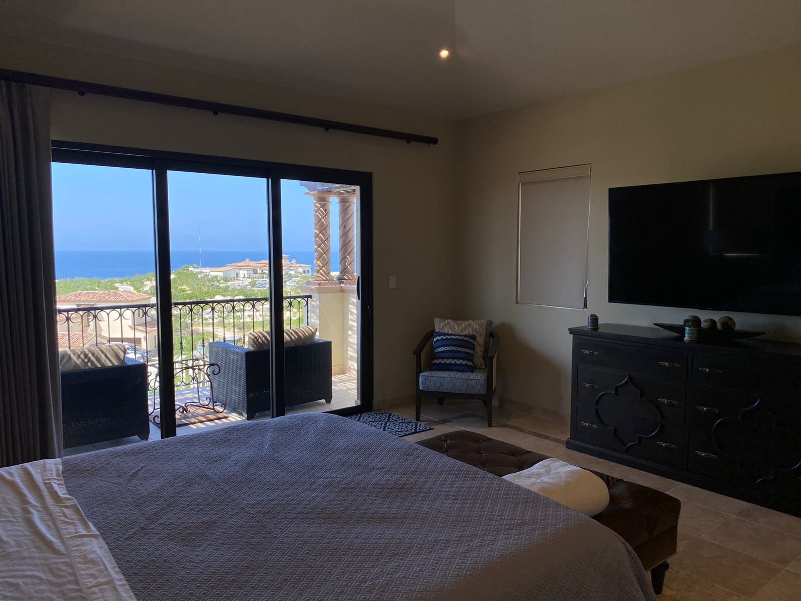 Room with large sliding glass door opening to balcony with outdoor seating, overlooking rooftops, trees, and ocean in the distance.