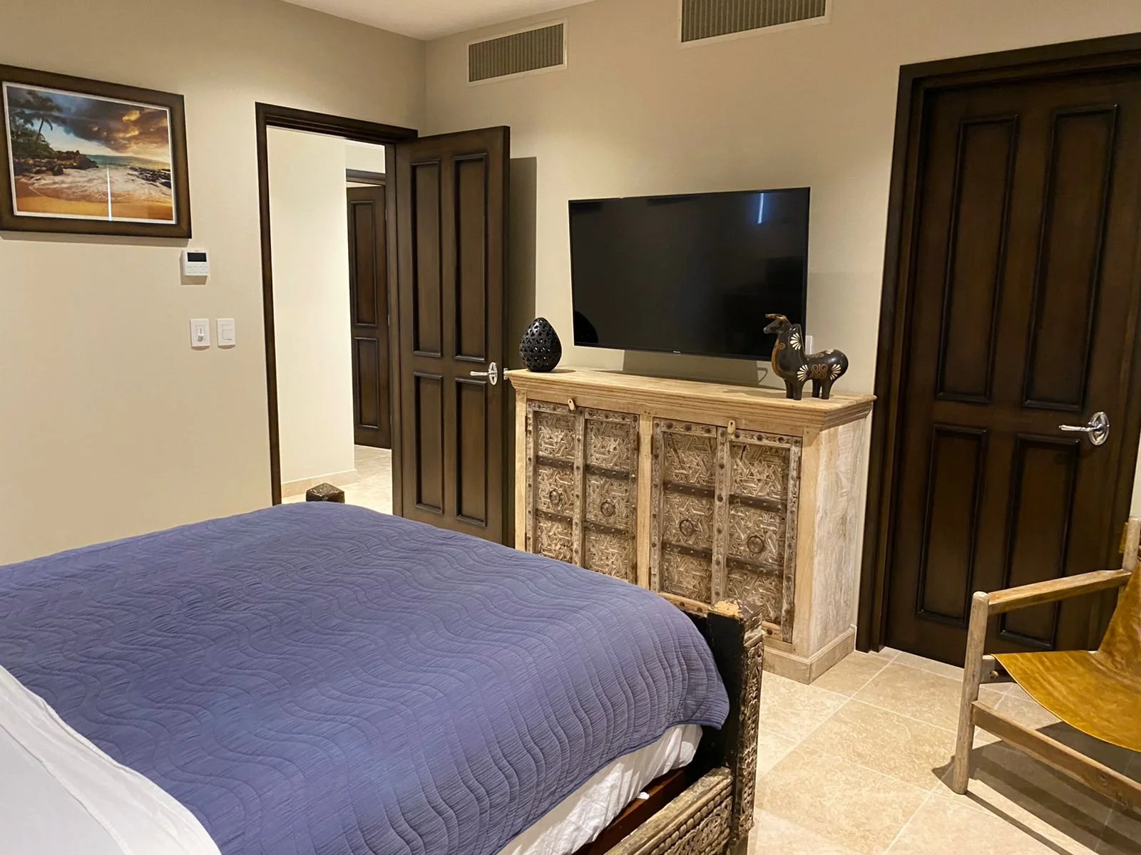 A bedroom with a bed covered in a purple quilt, dark wooden doors, a wooden dresser with intricate carvings, a flat-screen TV on top, and decorative items including a black vase and animal figurine. There is a picture of a beach scene on the wall and
