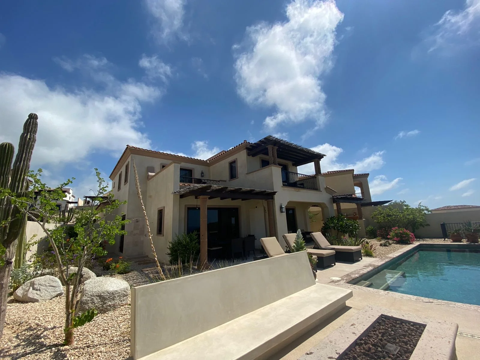 A modern two-story house with a swimming pool in the backyard, outdoor lounge chairs, desert plants, and a bright blue sky with scattered clouds.