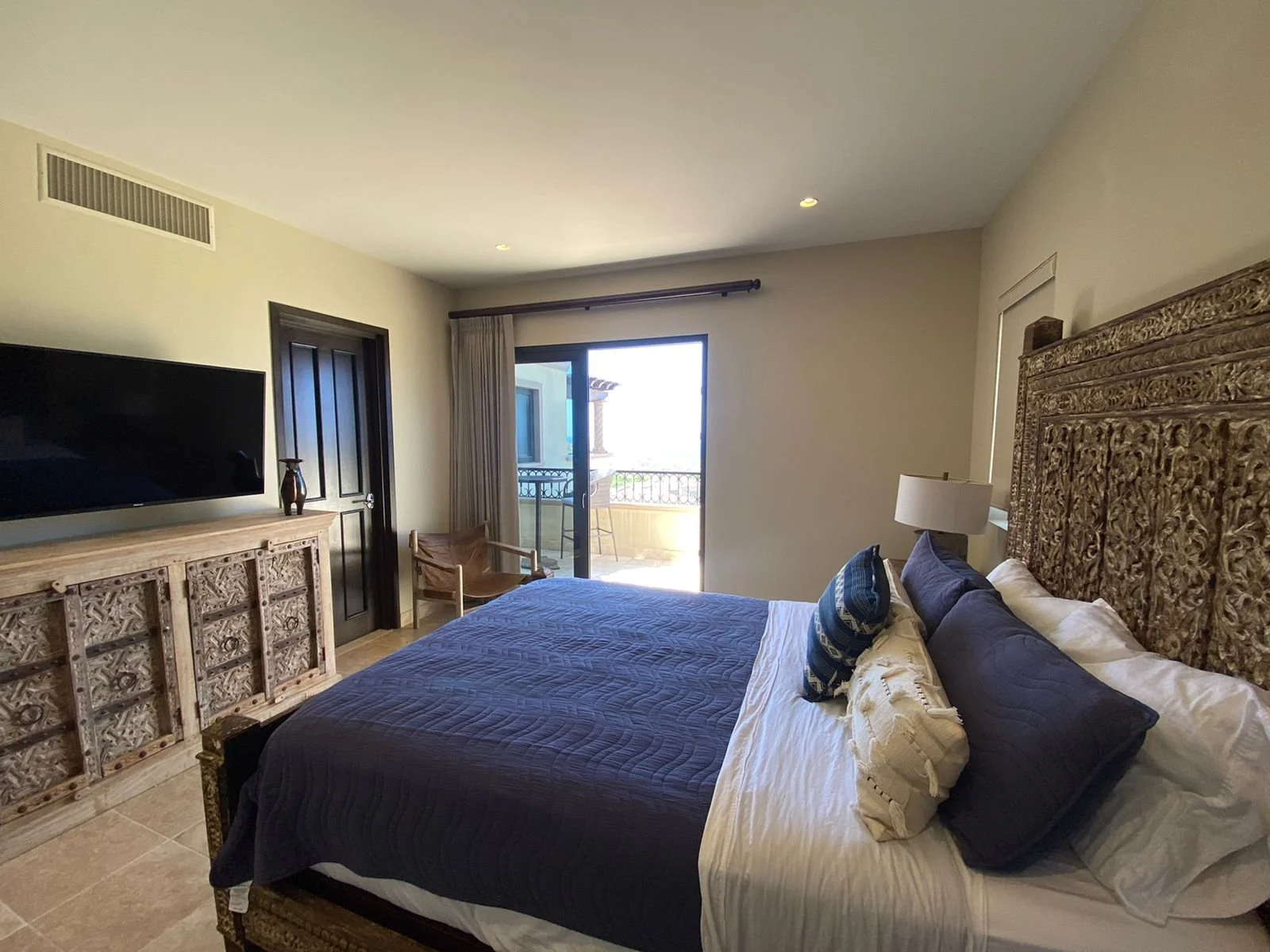 A bedroom with a large carved wooden headboard, a bed with navy and white bedding, a flat-screen TV on a carved wooden dresser, a chair near a sliding glass door leading to a balcony, and a window with curtains.
