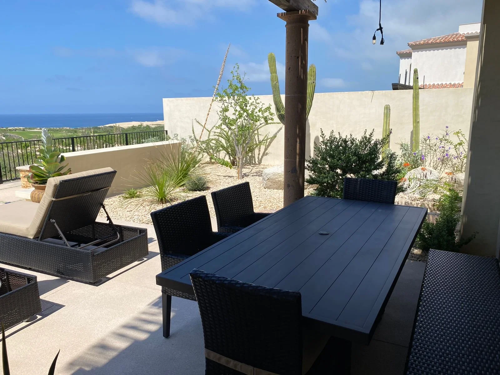 Outdoor patio with a black table and multiple black chairs, desert plants including cacti and succulents, view of ocean and horizon in the background.