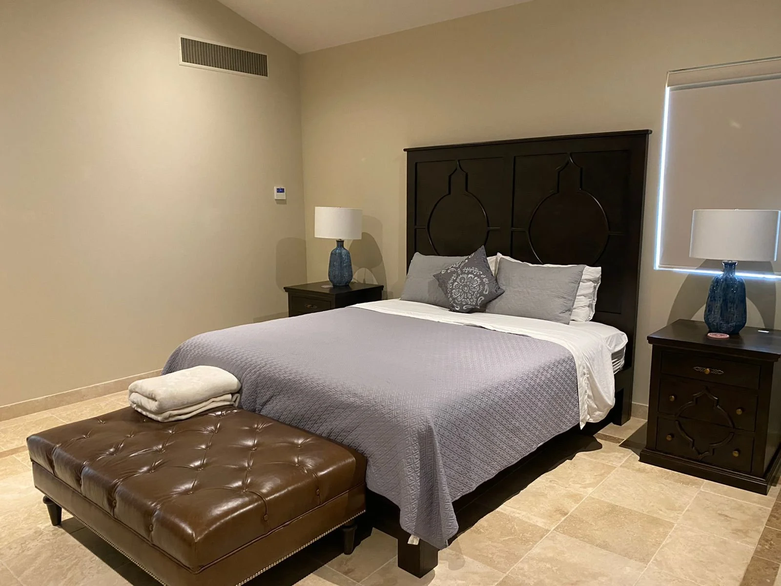A bedroom with a large bed, gray quilt, and decorative pillows, flanked by dark wood nightstands with blue lamps, a brown leather bench at the foot of the bed, and beige tiled flooring.
