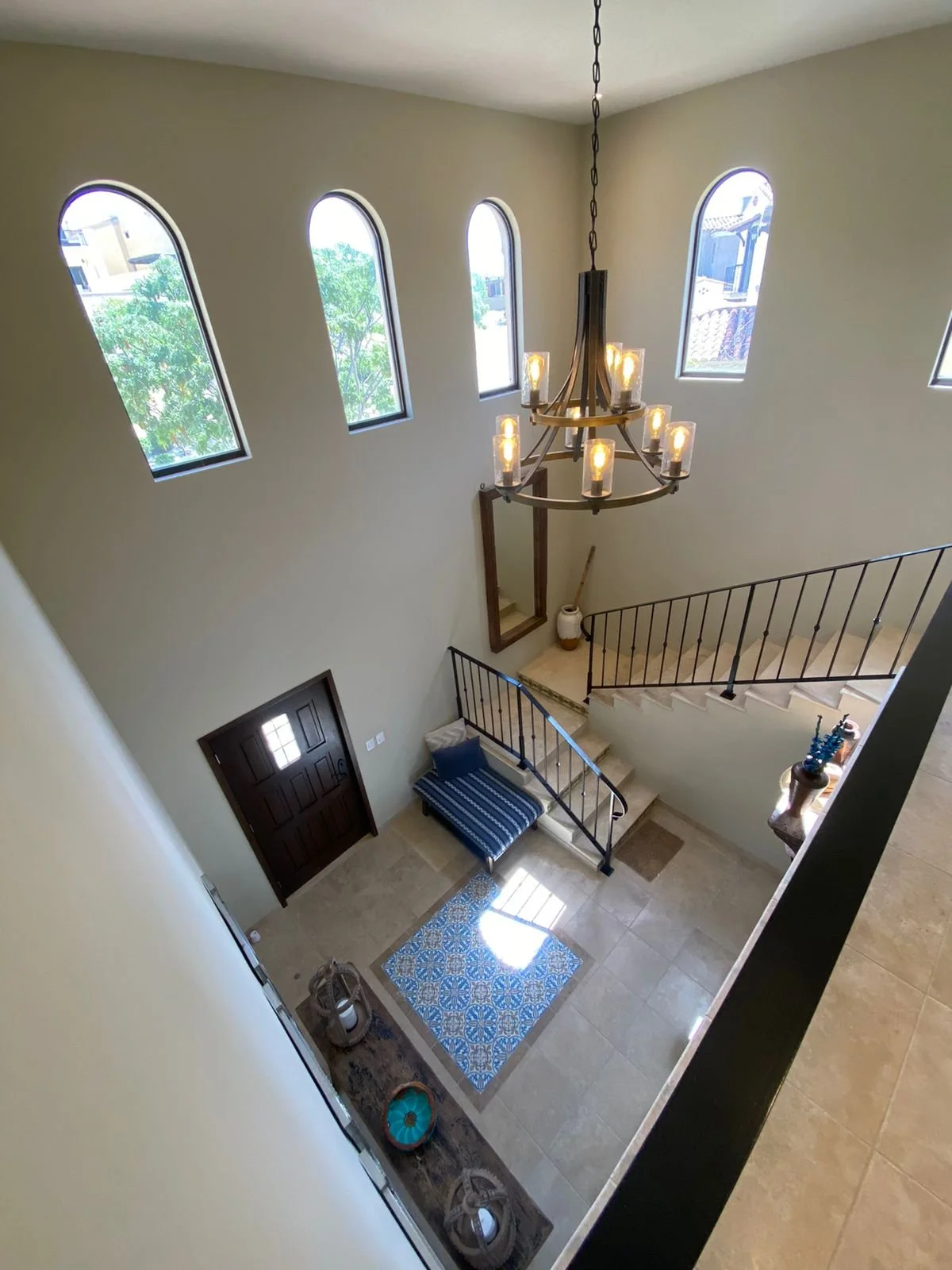 A view of a spacious foyer from above showing a dark wooden front door, tiled flooring, a blue patterned rug, a bench with striped fabric, a large chandelier, and high arched windows letting in natural light.