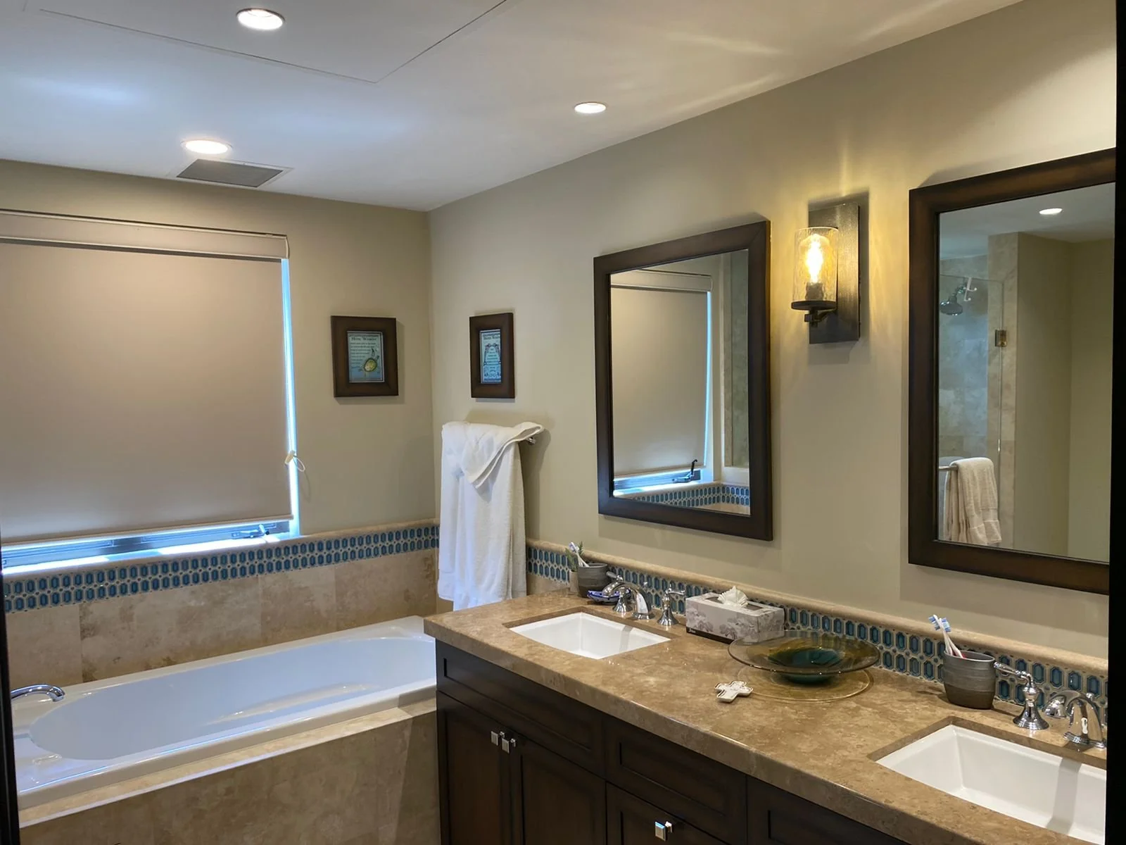 A modern bathroom with a double vanity featuring a beige marble countertop, two sinks, and dark wood cabinets. Above the sinks are two rectangular mirrors with dark wood frames. The room has beige tiled walls with a decorative blue tile border and a 