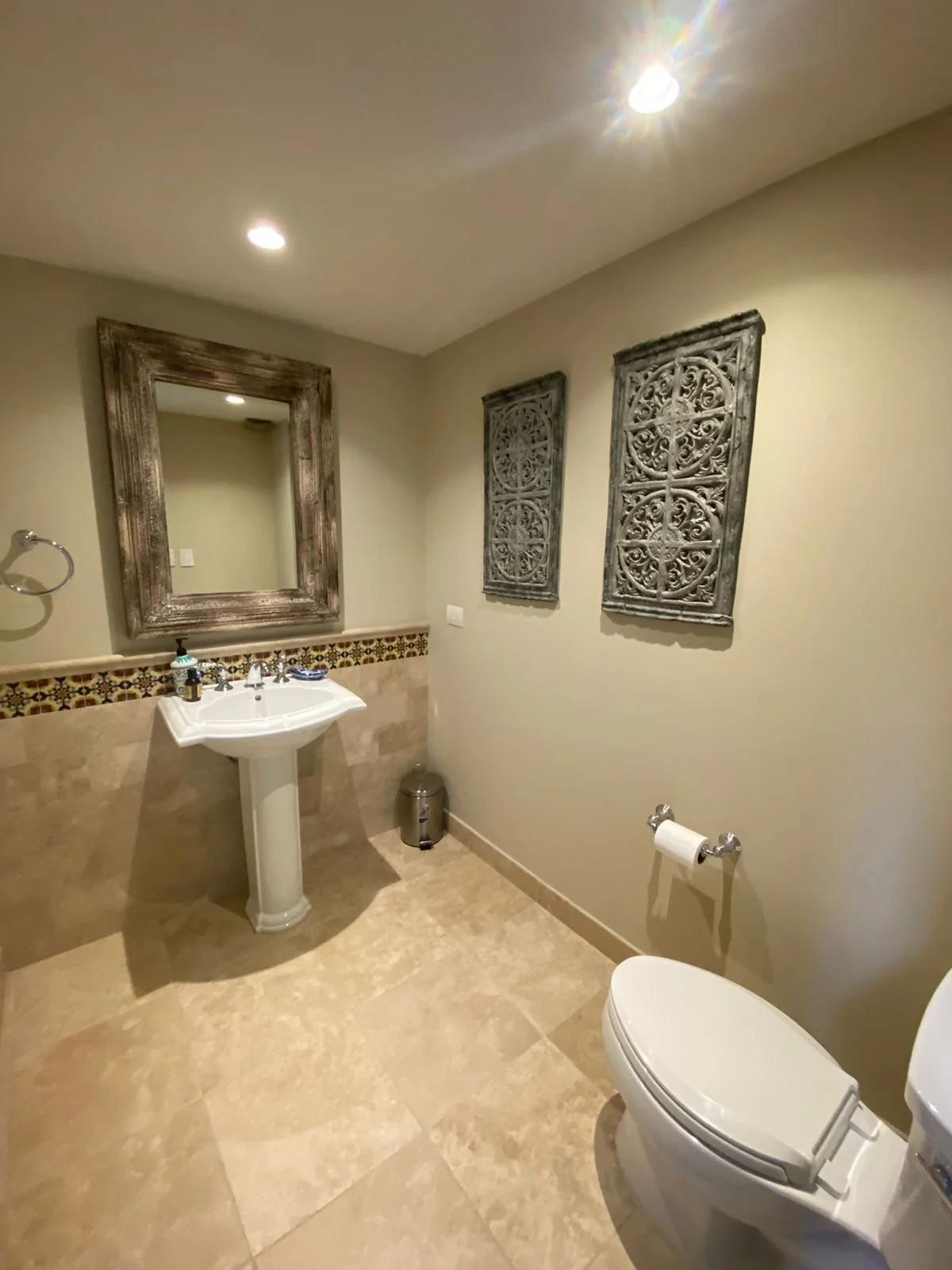 A bathroom with beige tile flooring, a white toilet, a pedestal sink with soap dispenser, a large framed mirror above the sink, two decorative wall hangings, a small trash bin, and recessed ceiling lights.