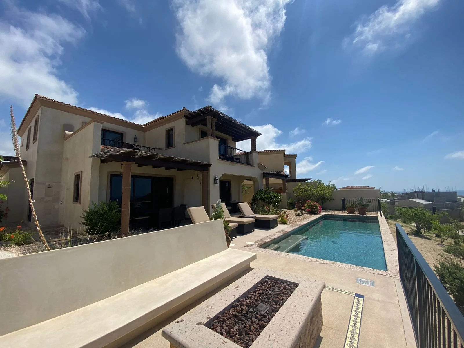 A luxurious two-story house with a swimming pool in the backyard, surrounded by patio furniture and plants, under a bright blue sky with scattered clouds.