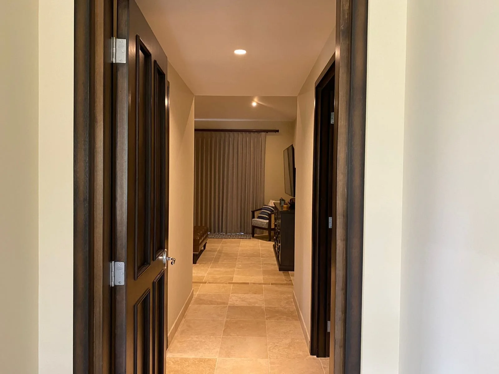 View of a hallway with beige tiled floor, beige walls, dark wooden door frames, and a ceiling with recessed lighting, leading to a living room area with a curtain, a TV on a stand, and a chair.