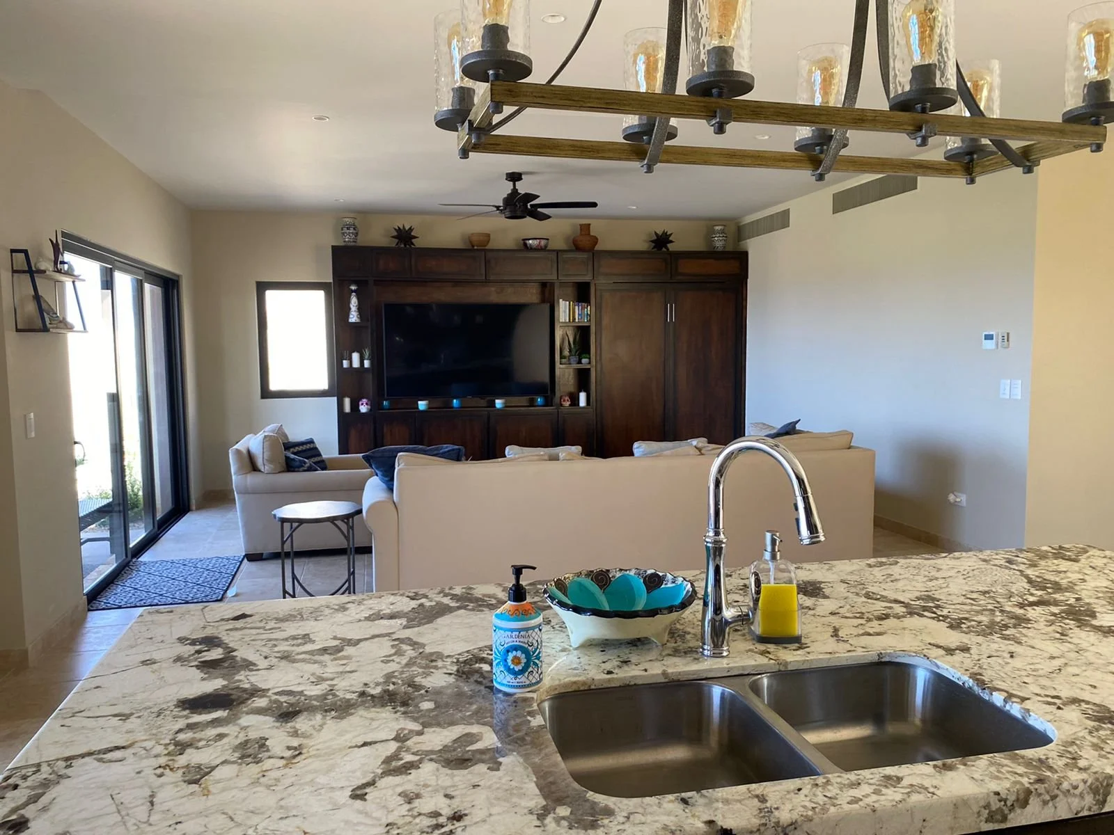 View from a kitchen island with a marble countertop featuring a soap dispenser, a bowl, and a hand sanitizer, looking out to a living room with beige sofas, a dark wood entertainment center with a large TV, and sliding glass doors.