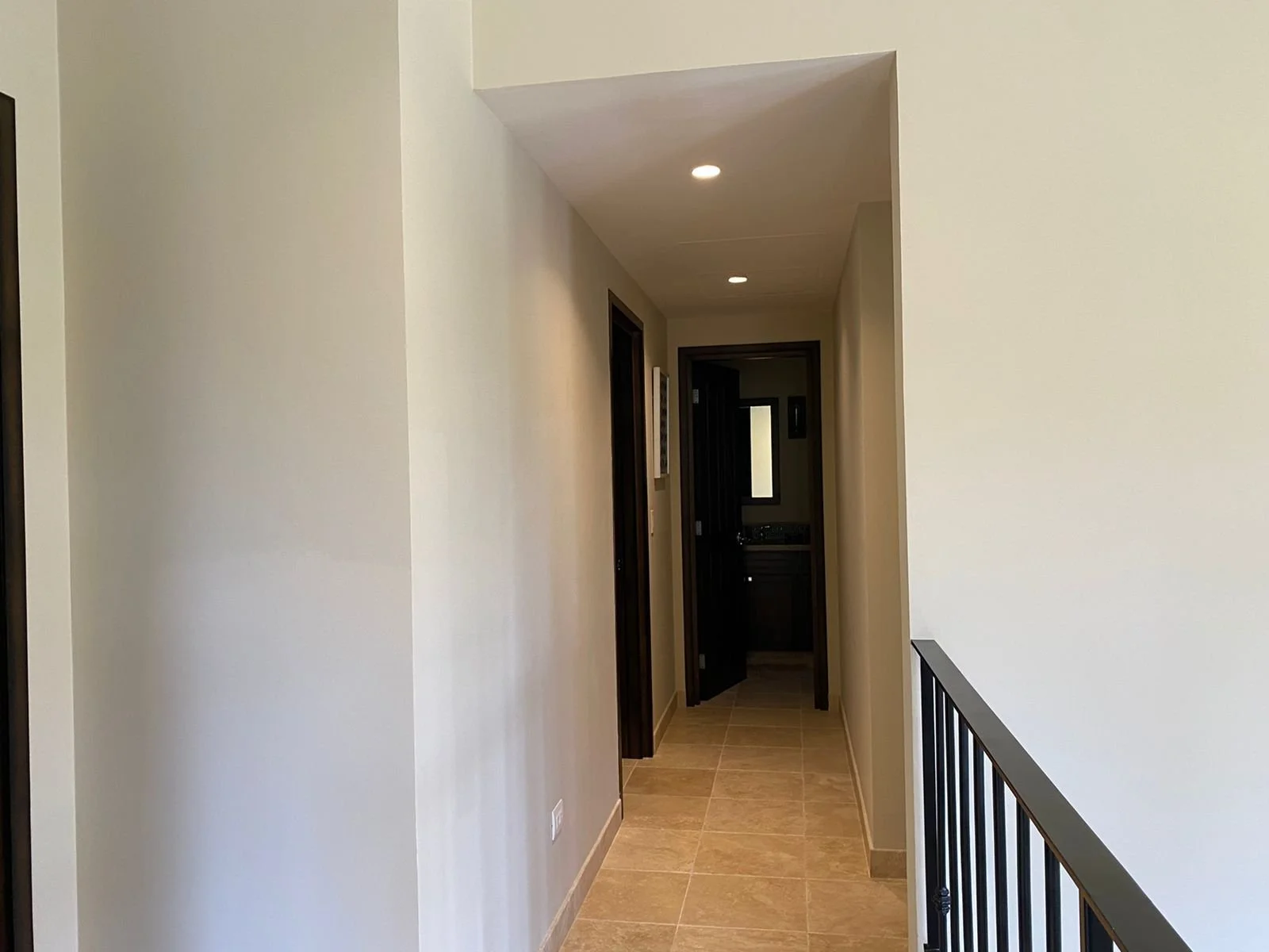 Hallway leading to a room with a window and dark door, beige tiled floor, cream-colored walls, and ceiling lights.