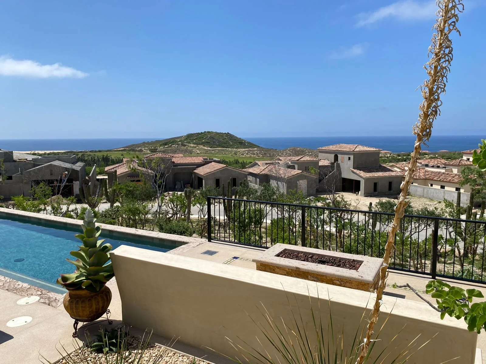 View of a suburban neighborhood with desert-style houses, a swimming pool in the foreground, and the ocean in the distance. Clear blue sky with a few clouds.
