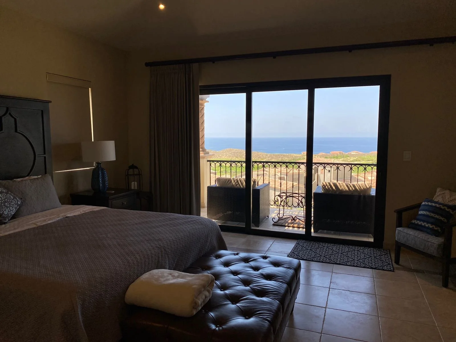 A hotel room with a large bed, nightstand, and table lamp, opening to a balcony with patio chairs, overlooking an ocean view.
