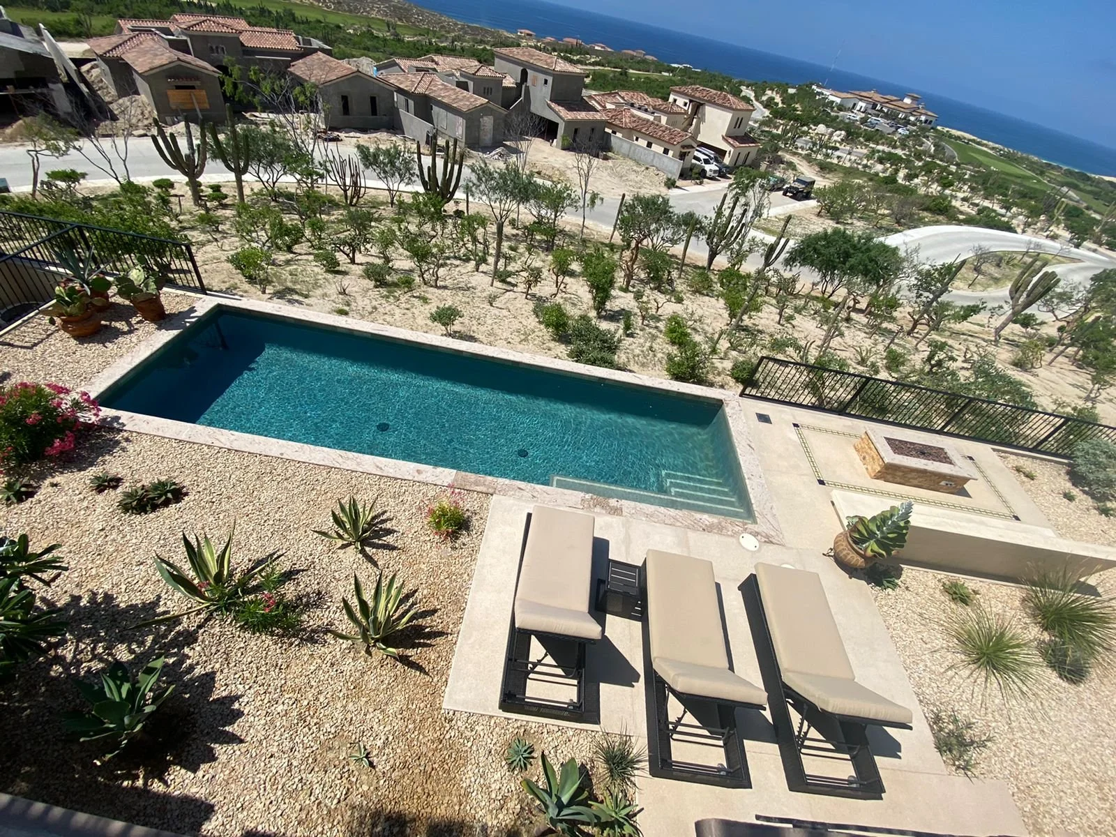 A backyard with a swimming pool, lounge chairs, desert plants, and a view of a neighborhood with houses, trees, and the ocean in the background.