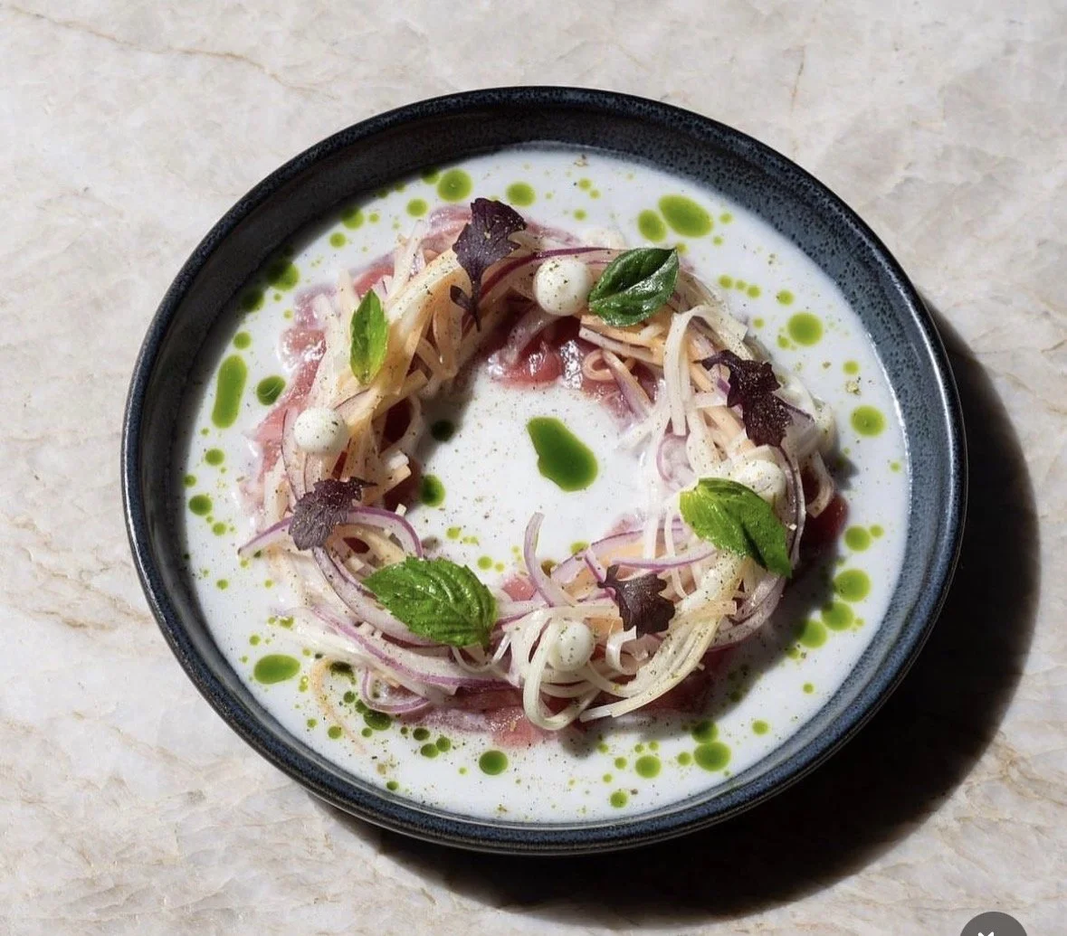 A dish of cold pasta salad in a black bowl, garnished with green herbs, with a white creamy sauce and green oil droplets on top.