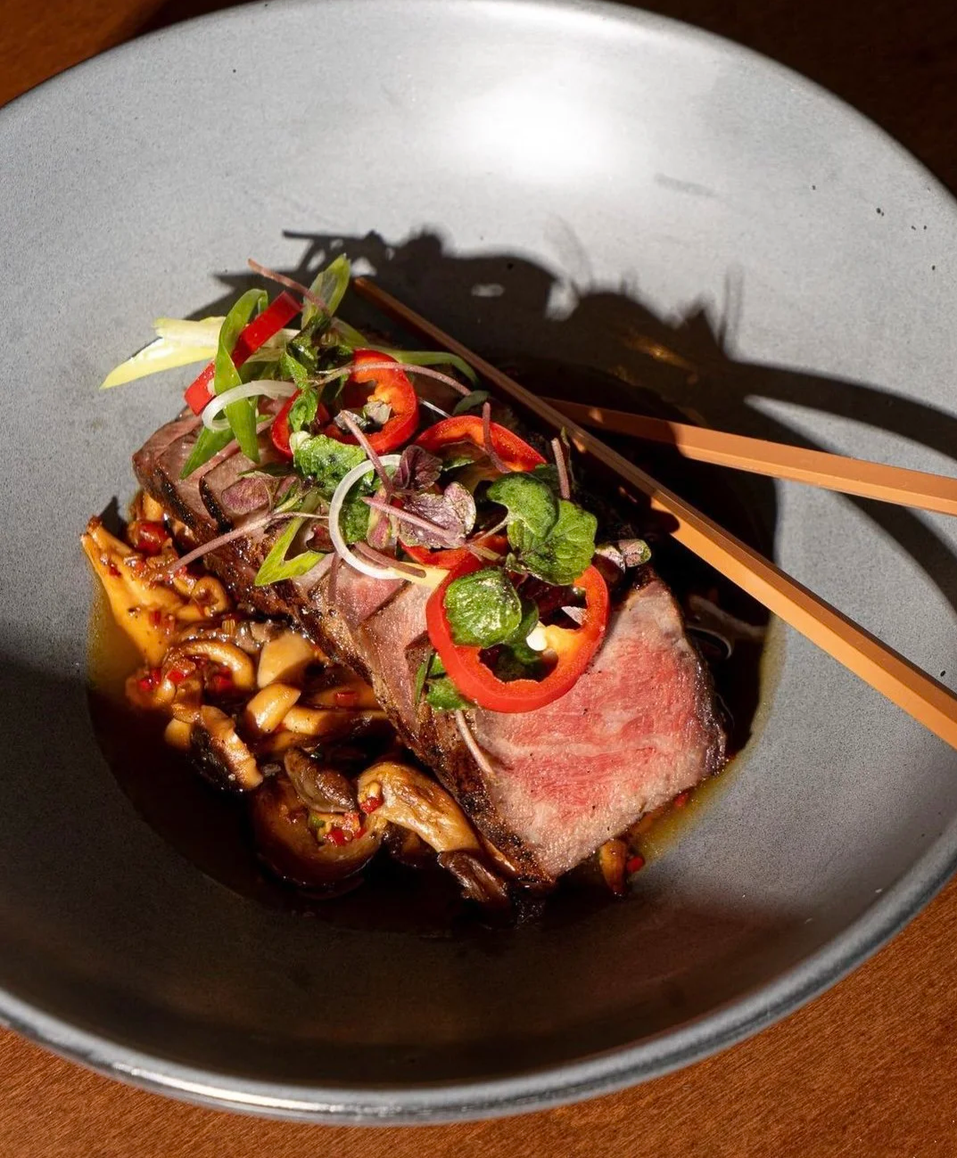 A plate with a cooked steak topped with sliced red chili peppers, garnished with microgreens and herbs, with a side of sautéed mushrooms and a sauce or oil drizzle.