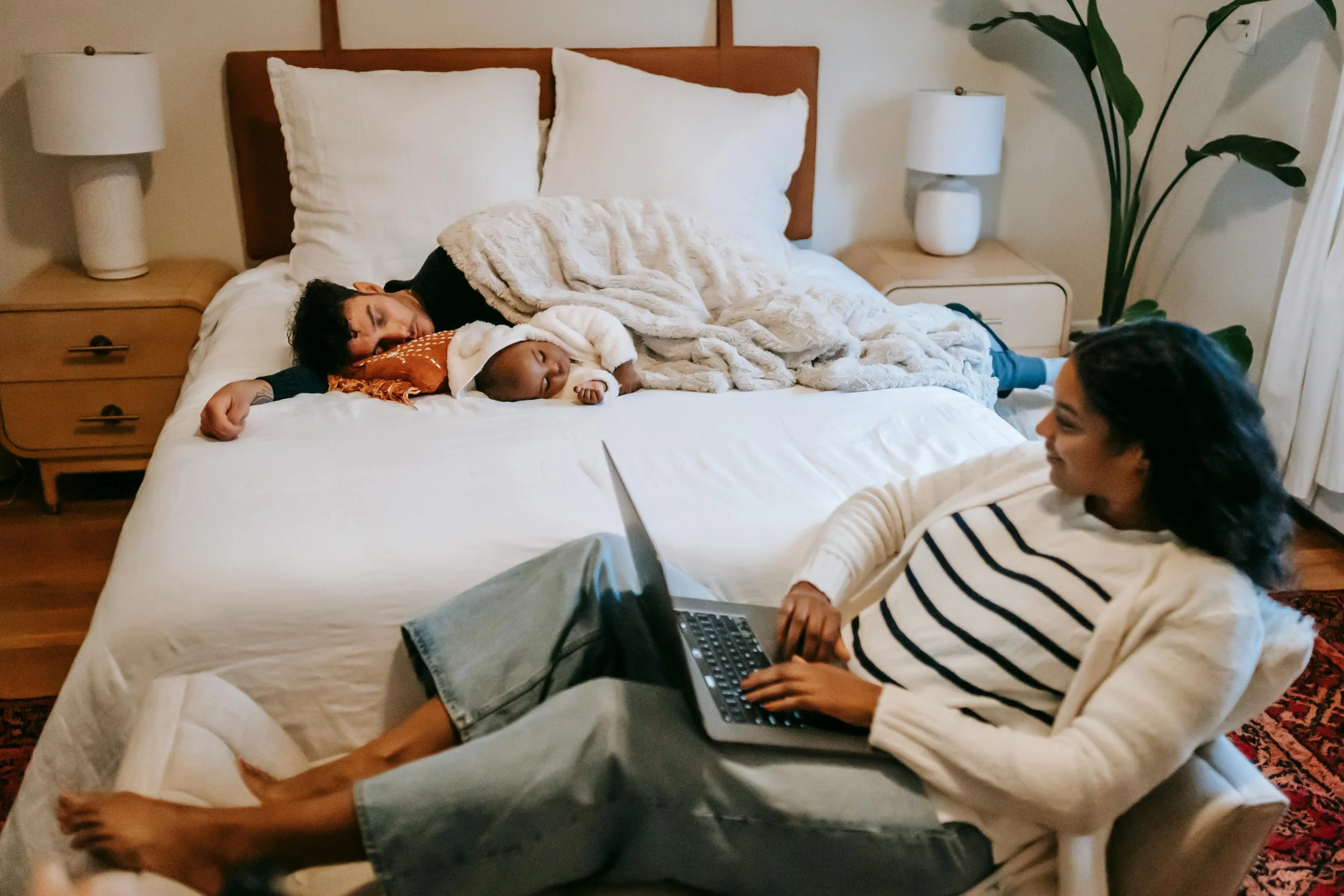 Woman sitting with a laptop while her partner and baby sleep, representing balancing responsibilities during a life transition.