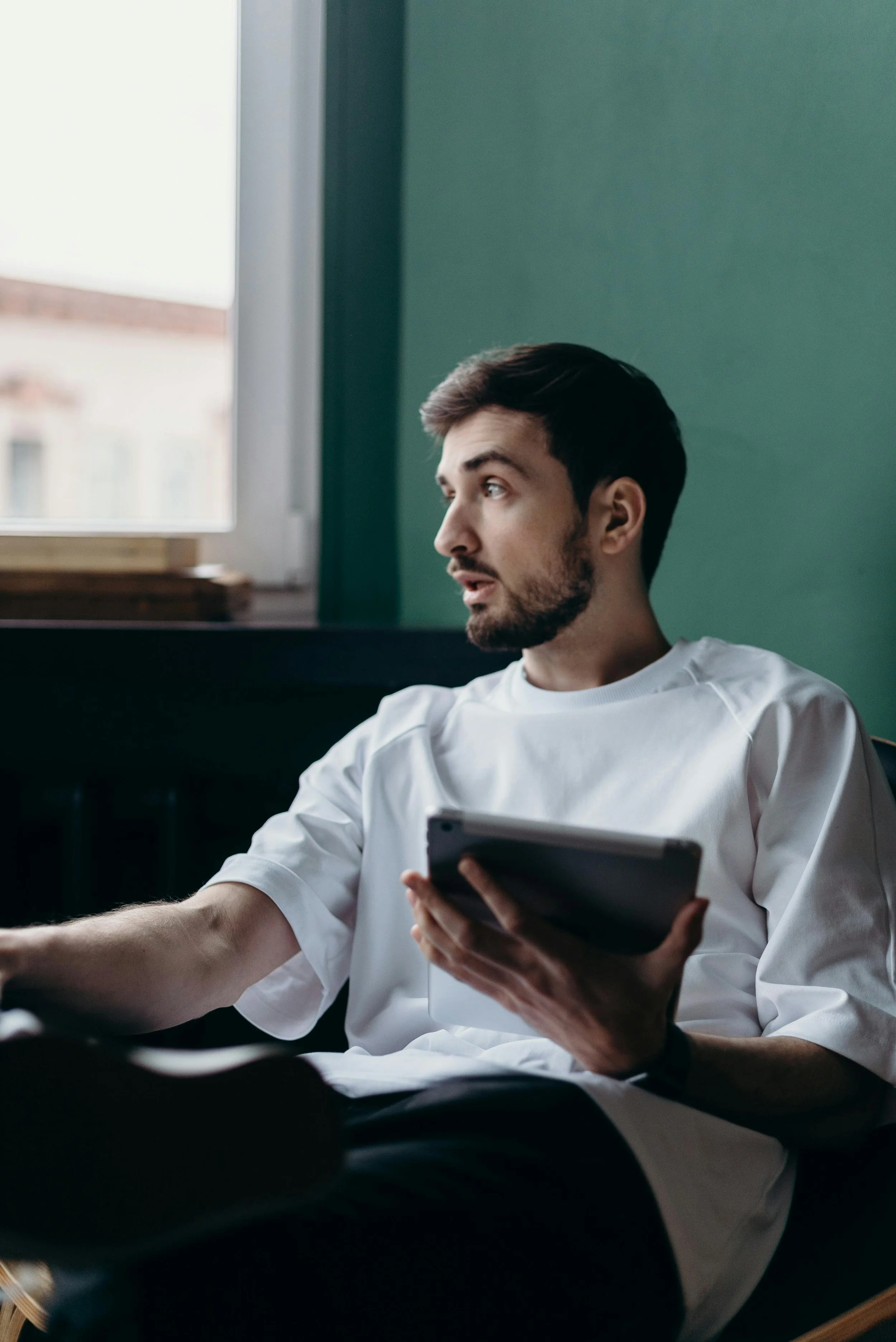 A man sitting on a couch, looking thoughtfully out the window while holding a tablet — suggesting reflection, stress, or quiet overwhelm.