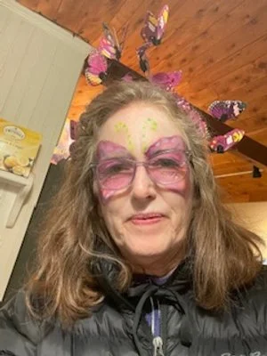 Woman with butterfly face paint, pink glasses, and butterfly headband in a cozy room with wood-paneled ceiling.