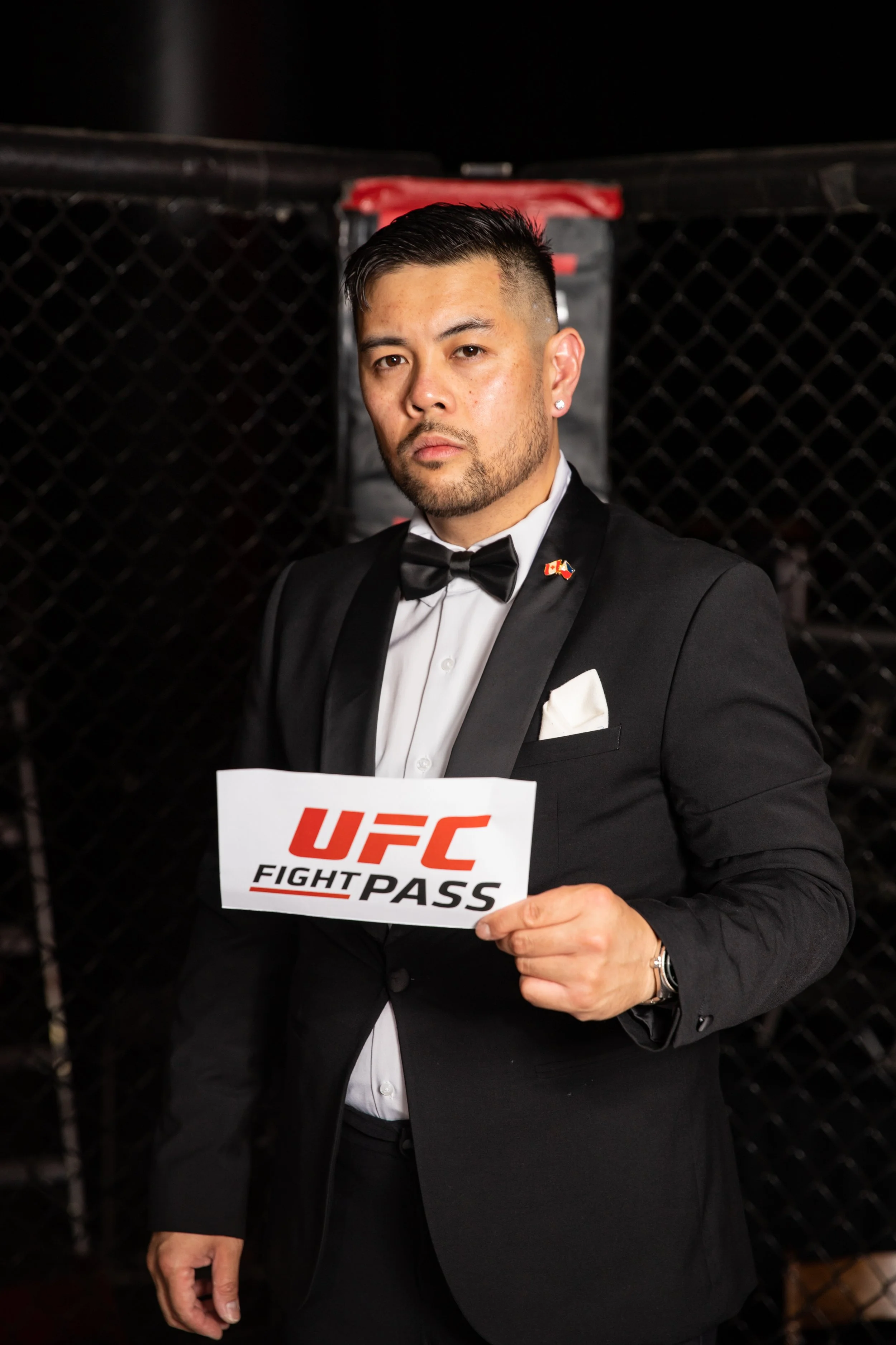 Ryan Ventura dressed in a black tuxedo with a bow tie and white dress shirt, holding a UFC Fight Pass sign, standing in front of a black cage.