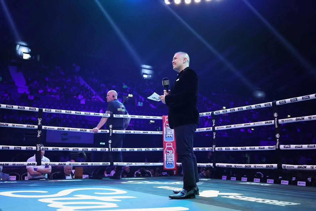 A boxing ring in a stadium with Ryan Ventura in a black suit holding a microphone and notes, standing in the ring, with two referees in the background, and spectators in the dark audience area.
