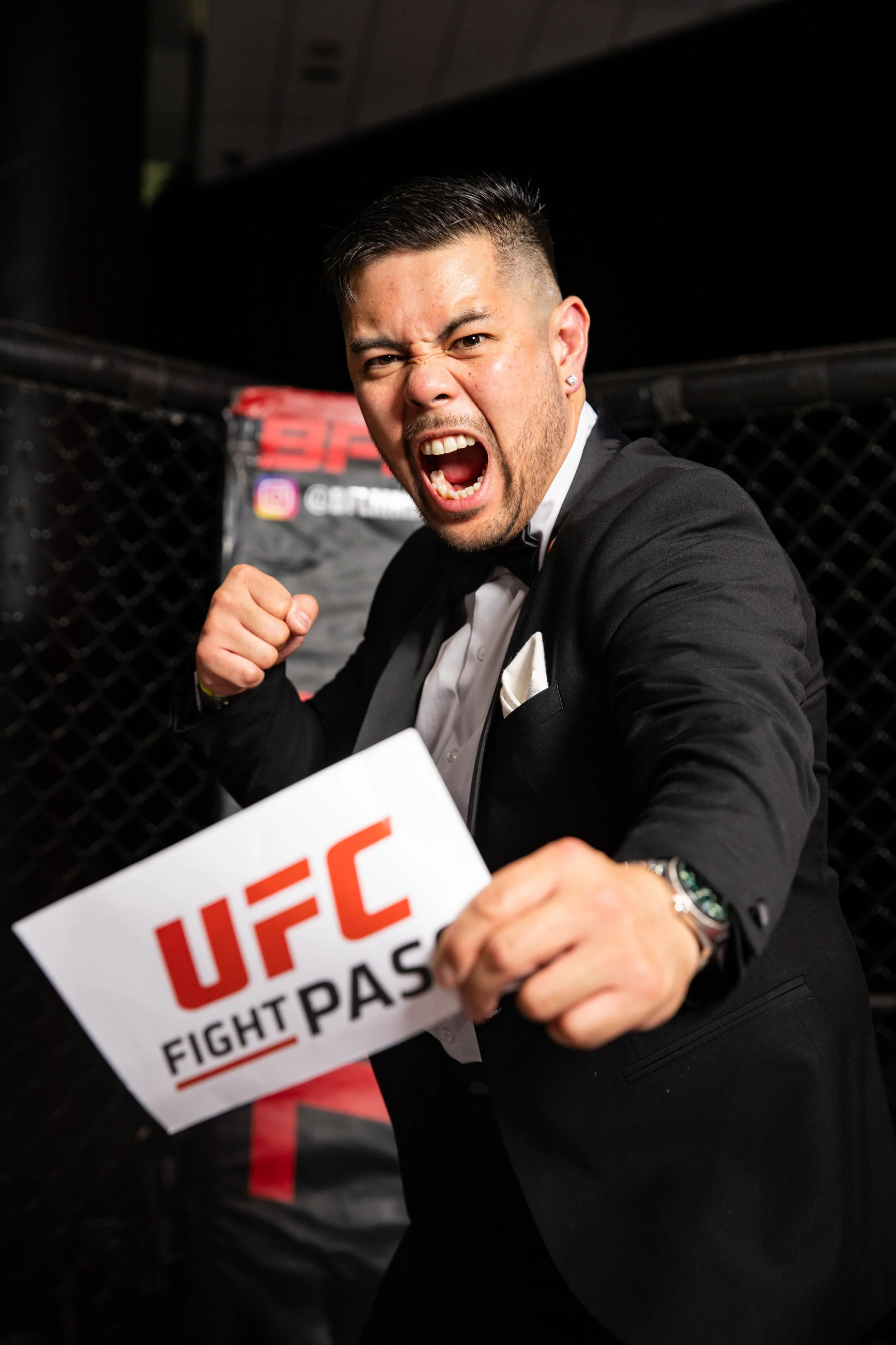 Ryan Ventura in a tuxedo making a fighting pose, holding a UFC fight pass sign, inside an octagon cage.