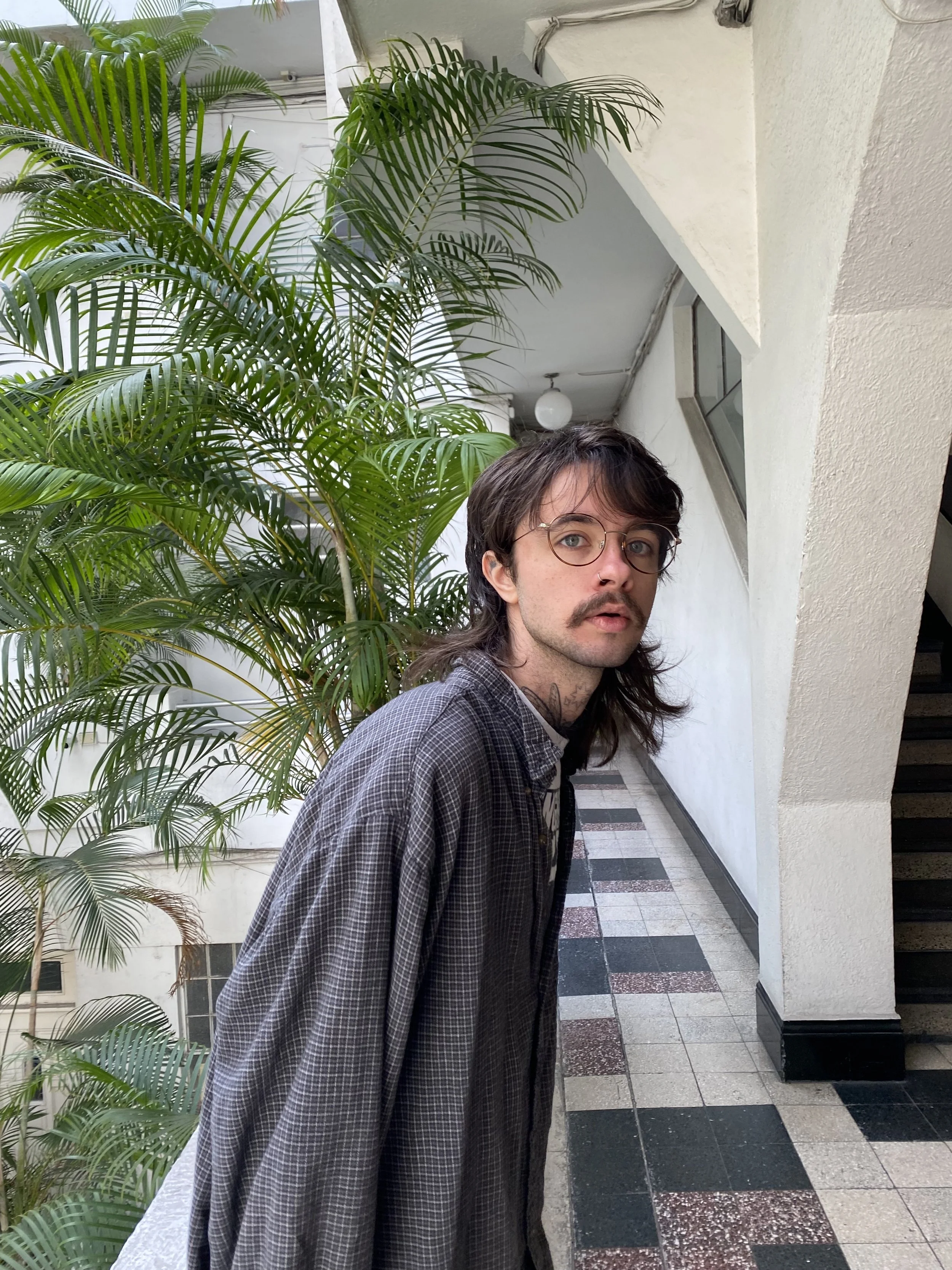 A young man with brown hair, glasses, and a mustache standing in a hallway next to a large green plant, wearing a loose checkered shirt.