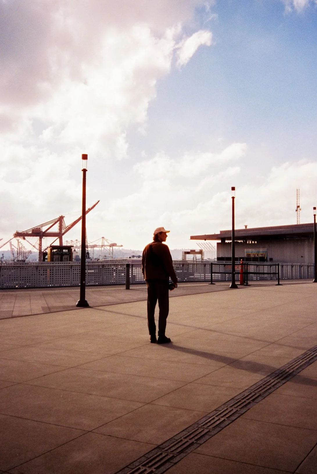 A person stands on a spacious, empty dock or promenade area, with industrial cranes and buildings in the background under a partly cloudy sky.