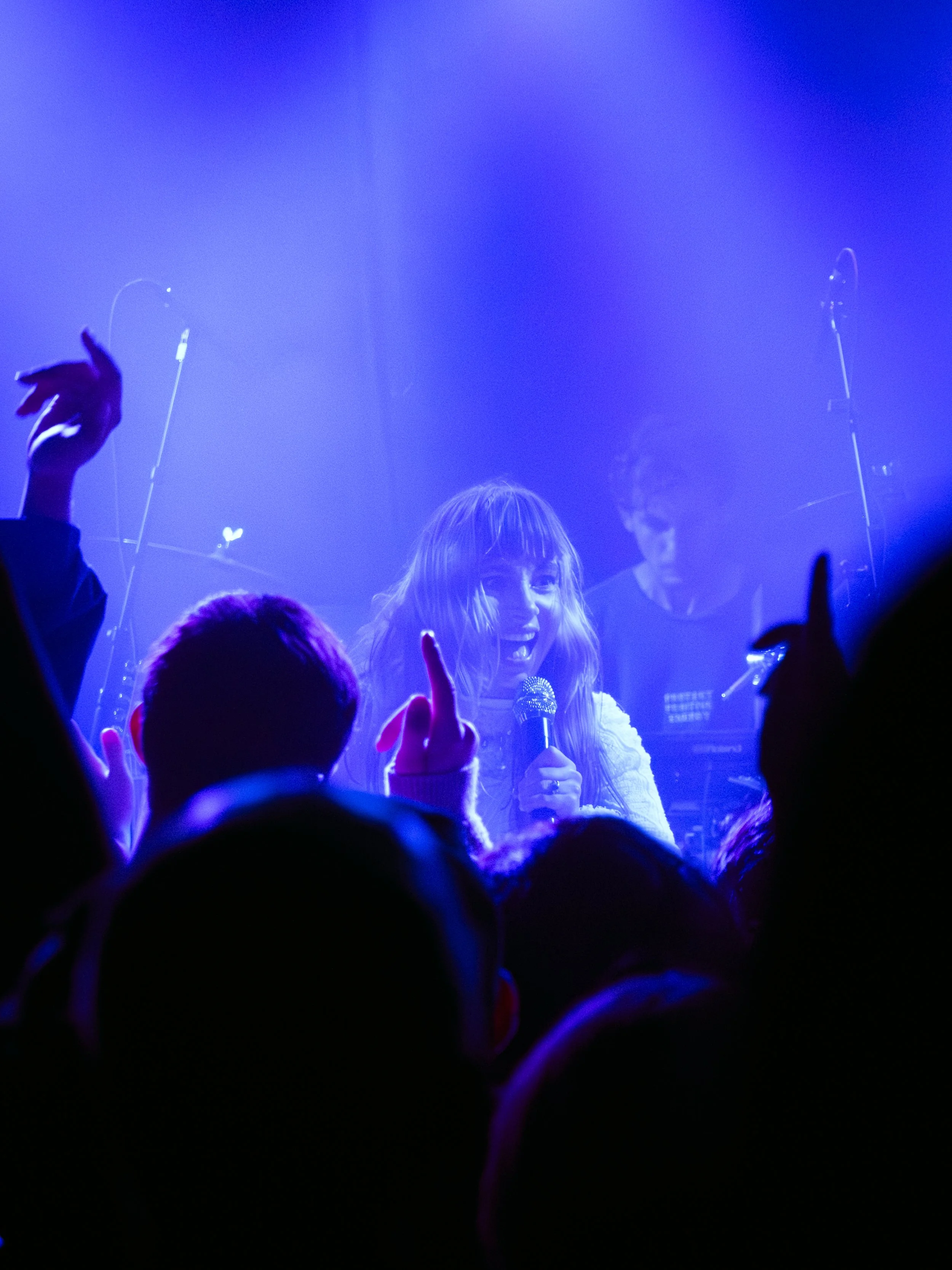 Female singer holding microphone performing on stage with audience in front, blue lighting, and a male musician playing keyboard in background.