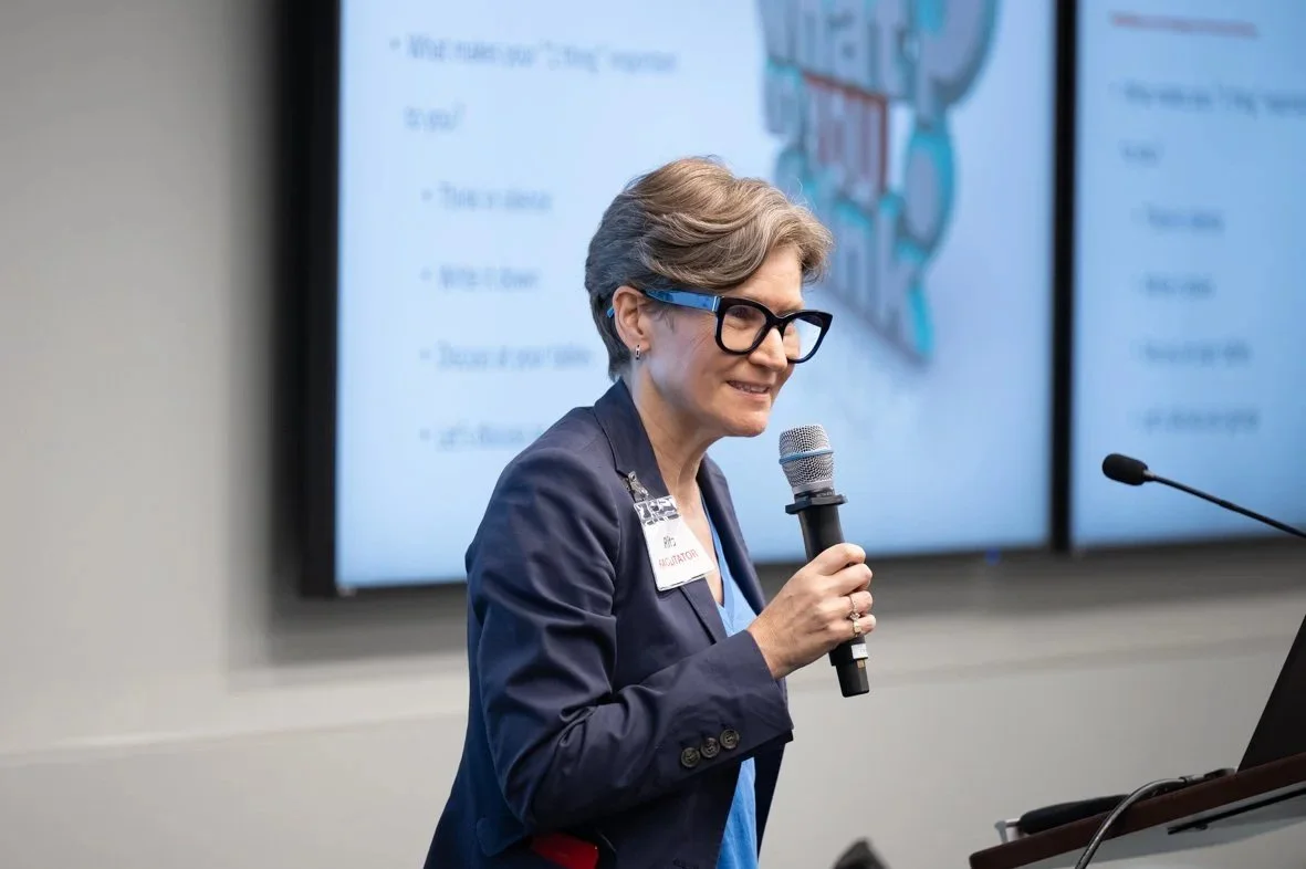 A woman with short gray hair, glasses, and a navy blazer speaking into a microphone during a presentation in front of a large screen displaying slides.