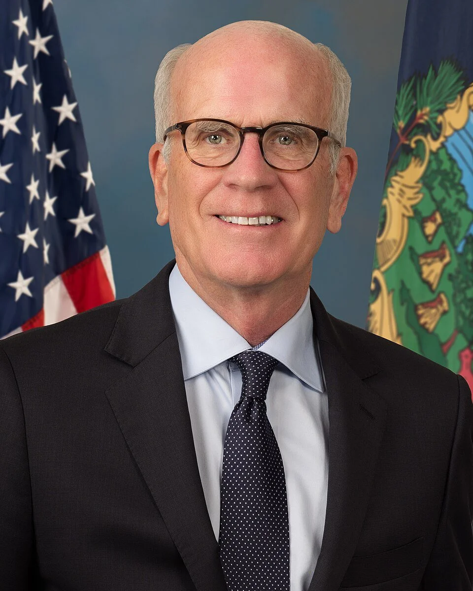 An older man with glasses, white hair, wearing a suit, tie, and white shirt, standing in front of American and state flags.