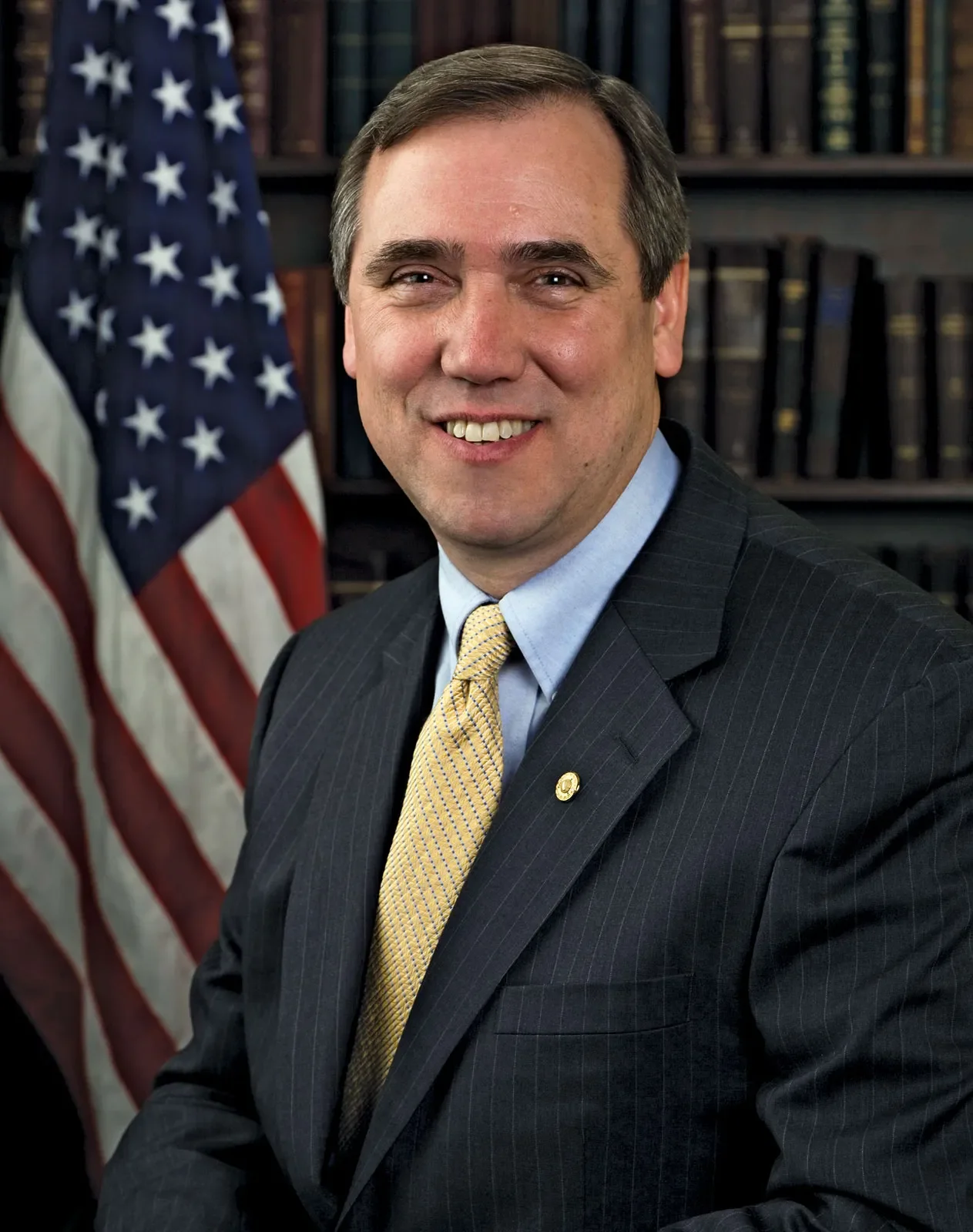 A man in a dark suit with a yellow tie, smiling, sitting in front of an American flag and a bookshelf filled with books.