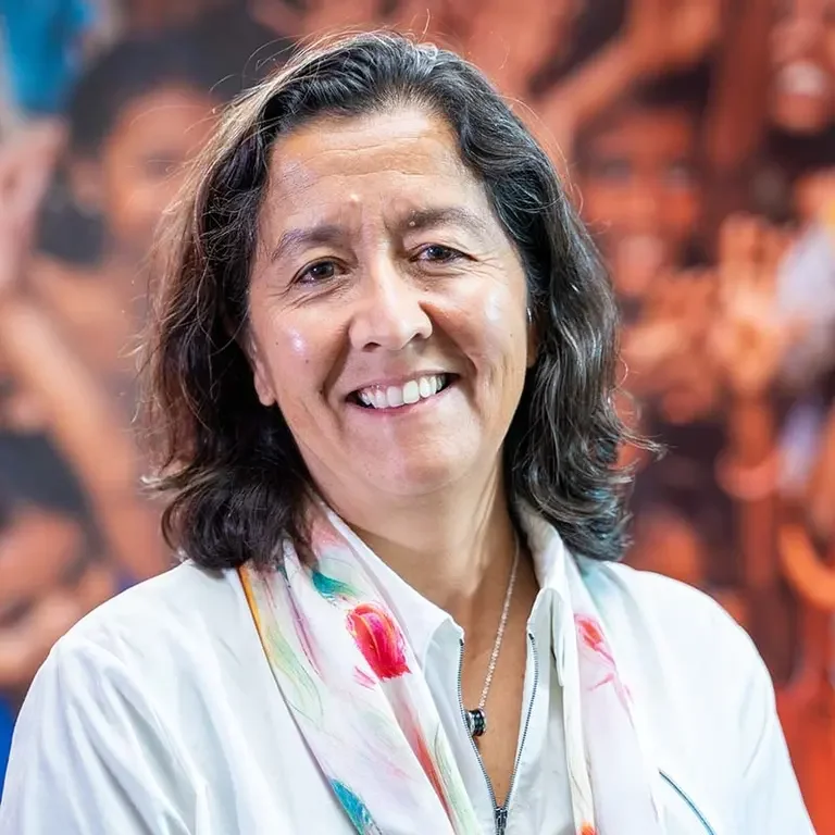 A woman with medium-length dark hair smiling, wearing a white shirt and a colorful scarf, standing in front of a blurred background of orange and brown hues.
